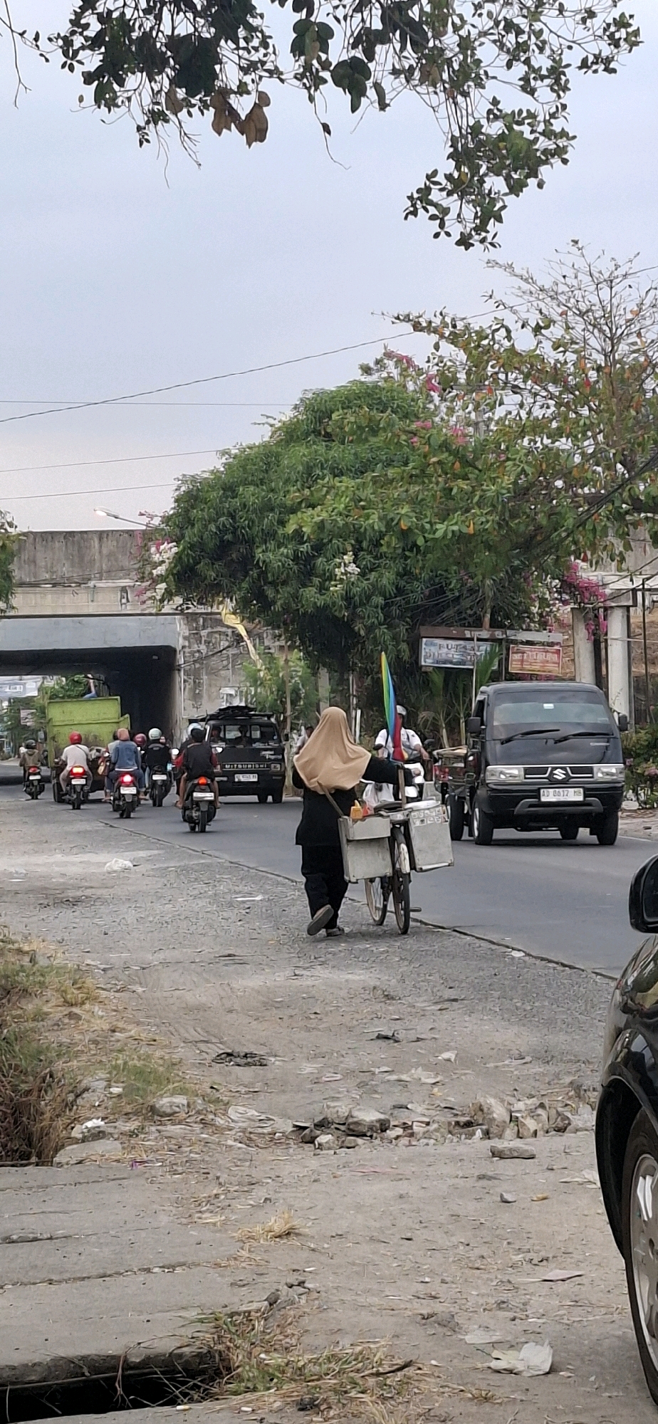 The image depicts a street scene in Desa Dibal, Indonesia, likely during daylight hours given the overcast sky. The foreground shows a rough, debris-strewn roadside with grass. A black car is partially visible on the far right. A woman wearing a tan hijab and black clothing walks away from the viewer, pushing a three-wheeled cart attached to a bicycle. The cart is silver and appears to hold containers. Behind her, a black Suzuki carry van with license plate "AD 0617 HB" is driving on the asphalt road. To the left of the van, a black Mitsubishi truck with a license plate that is partially visible is also on the road. Further left, several motorcycles are traveling in the same direction. One motorcycle rider wears a red helmet, another a blue helmet, and a third has a black helmet. In the background, a green dump truck is visible under an overpass. The overpass is constructed of concrete and appears to be part of a road or railway. Trees with green foliage and some pink flowers are prominent along the roadside and near the overpass. A rainbow-colored flag is attached to a pole on the right side of the image, near the Suzuki van. The overall environment suggests a busy street with moderate traffic and a mix of urban and natural elements.