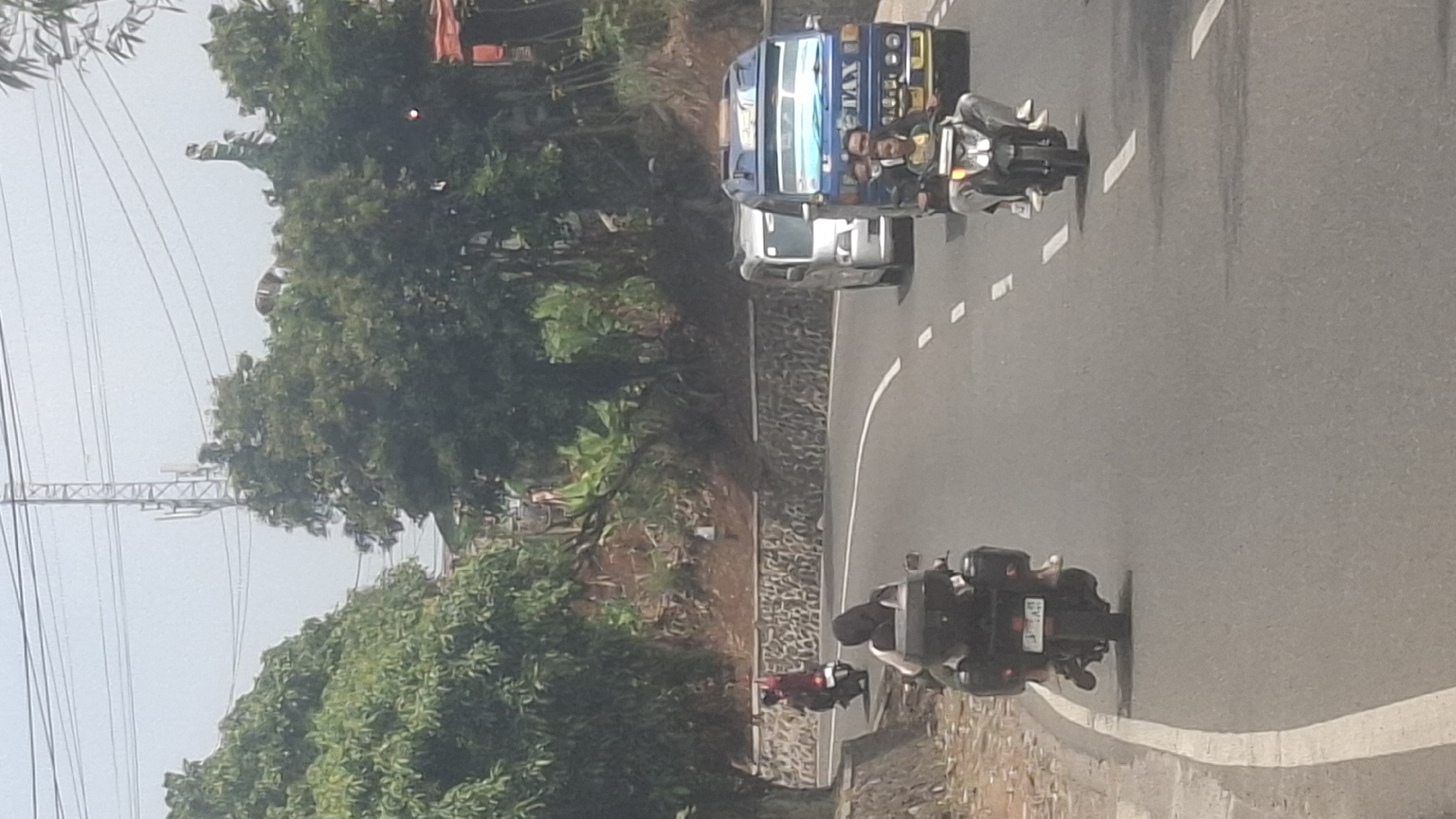In Sirnagalih, Indonesia, on a clear day, a paved road curves through a hilly, green landscape dotted with trees. Several vehicles are visible on the road. In the foreground, a motorcycle with two passengers, a driver and a pillion rider wearing a headscarf, travels away from the camera. The motorcycle has two black saddlebags. Further up the road, another motorcycle with a single rider, also wearing a headscarf, moves away from the camera. A light-colored van is positioned to the right of the road, followed by a blue vehicle that appears to be a small bus or van with "TAX" written on its side, also carrying passengers. The background features a stone-lined embankment and dense foliage, with a tall telecommunications tower visible in the distance against a hazy sky. Power lines traverse the upper portion of the image.
