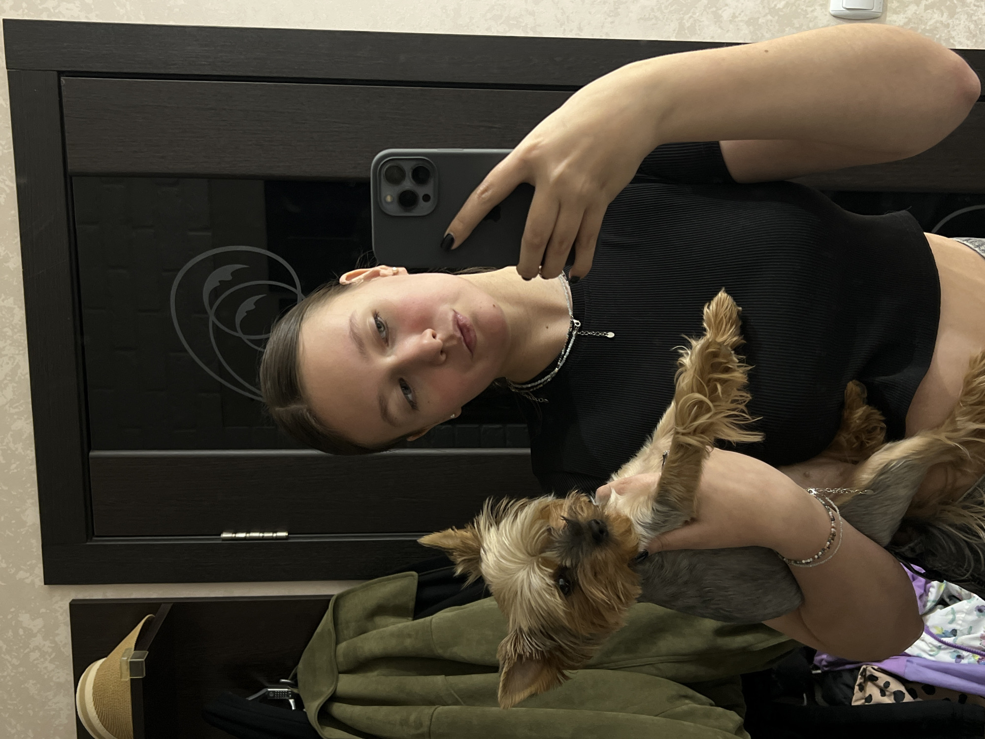 A young woman is holding a small dog and taking a selfie with her phone. She is wearing a black top and a silver necklace. The dog is a Yorkshire Terrier with a gray body and brown and blonde fur on its head and ears. The background shows a dark brown wardrobe, a light-colored wall, and a green jacket or coat. A hat is hanging on the wardrobe. The location is Chaikovsky, Russia.