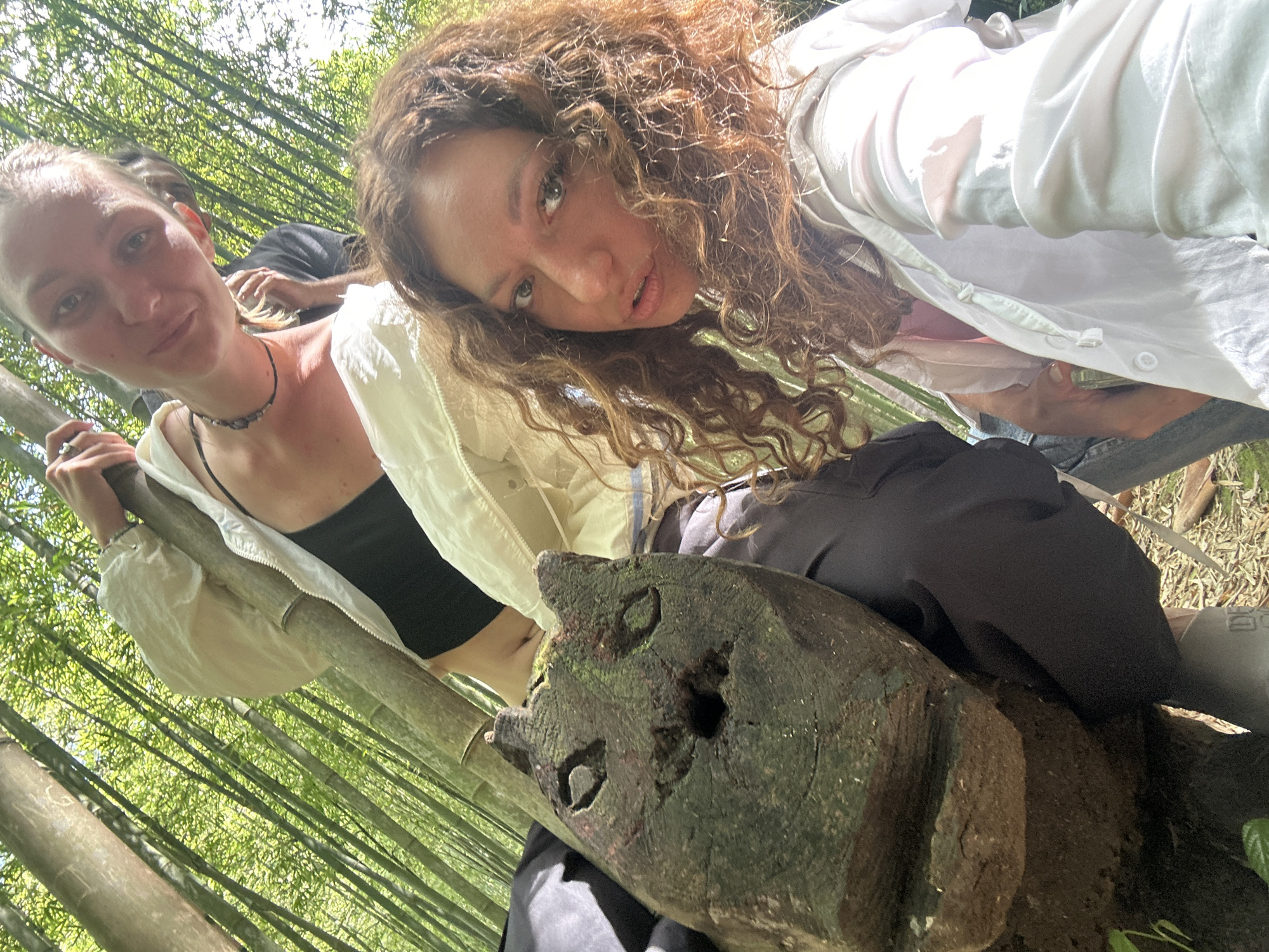 Two women pose for a selfie in a bamboo forest. The woman on the left has her hair pulled back and wears a black top, white jacket, and a necklace. The woman on the right has curly hair and wears a white shirt. Both are leaning over a piece of carved wood. The bamboo forest is dense. The photo appears to have been taken in Púng Luông, Vietnam.