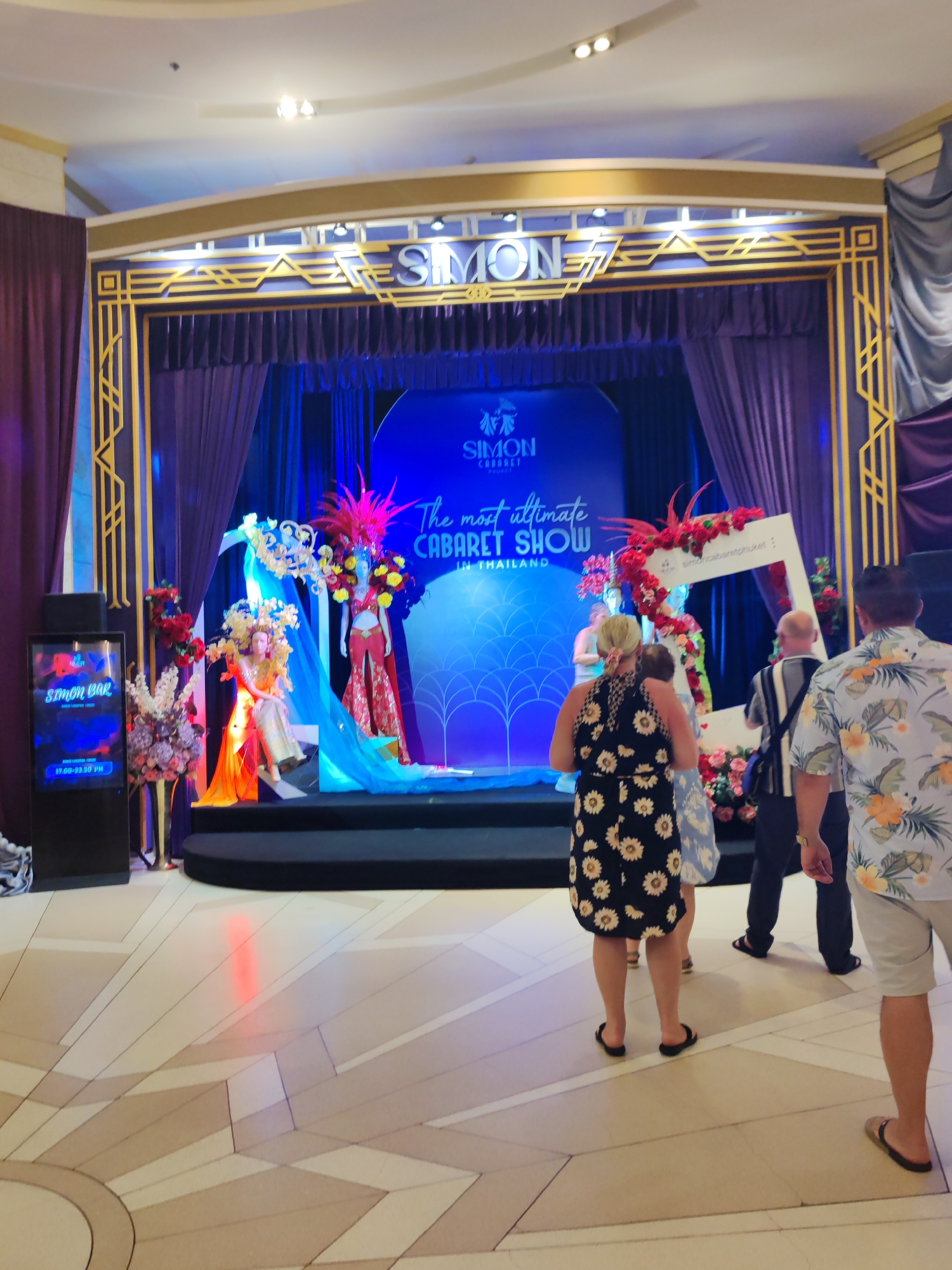 Here is a detailed description of the image:
The image shows a stage setup for the Simon Cabaret Show in Patong, Thailand. The stage is adorned with purple curtains and a decorative golden frame spelling out "SIMON". Mannequins in vibrant, elaborate cabaret costumes and headdresses are positioned on the stage.
Several people are standing in front of the stage. From left to right are an electronic sign that says SIMON BAR, 3 people standing towards the center, and one person to the right. The image appears to be taken in a shopping center, judging by the tiled floor and building structure.