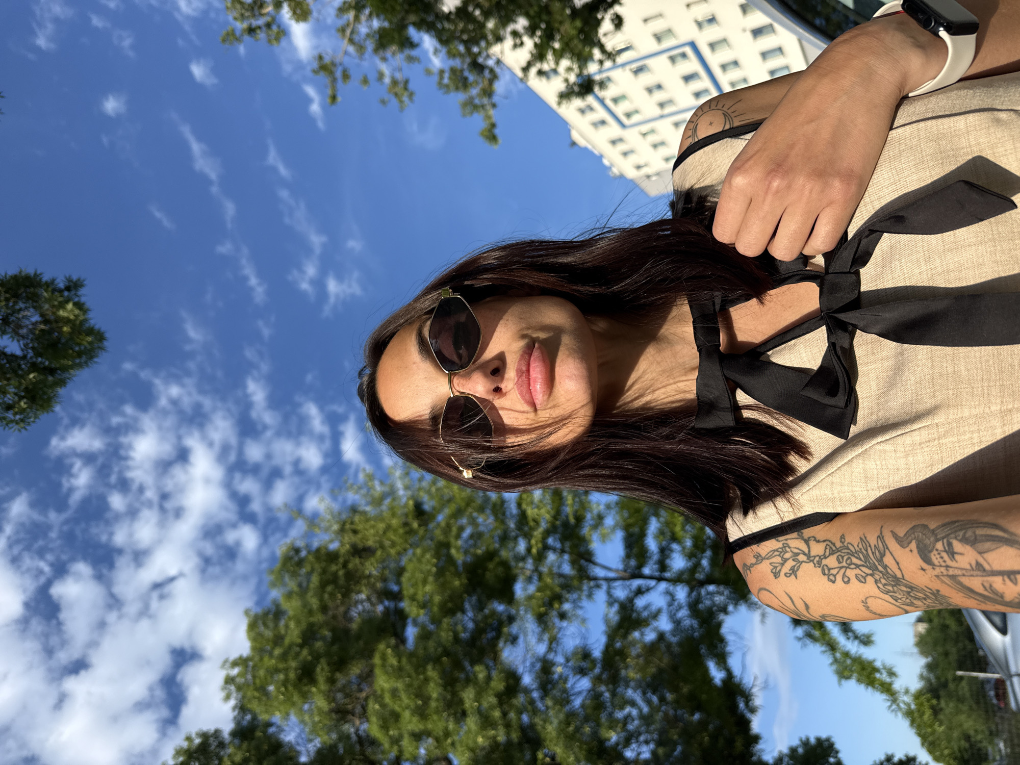 A vibrant outdoor portrait features a woman captured from a low angle, looking up towards the camera. The original image is rotated 90 degrees clockwise. She has long, dark hair flowing around her face and is wearing hexagonal-framed sunglasses. Her lips are slightly parted in a relaxed expression. She is dressed in a light beige or tan top adorned with black ribbon ties and bows around the neckline. Visible tattoos decorate her arms: a detailed botanical or organic design covers her left upper arm, and a sun-and-moon motif is present on her right shoulder. A white-banded Apple Watch is worn on her right wrist.
The background consists of a clear, bright blue sky with scattered white, wispy clouds. Green trees frame the bottom and sides of the shot, suggesting a natural, possibly park-like setting. In the upper right background, a light-colored building with repetitive square windows is visible against the sky. The scene is illuminated by strong, natural sunlight, indicating a sunny daytime. The woman appears to be posing casually for the photograph. No text is visible in the image.