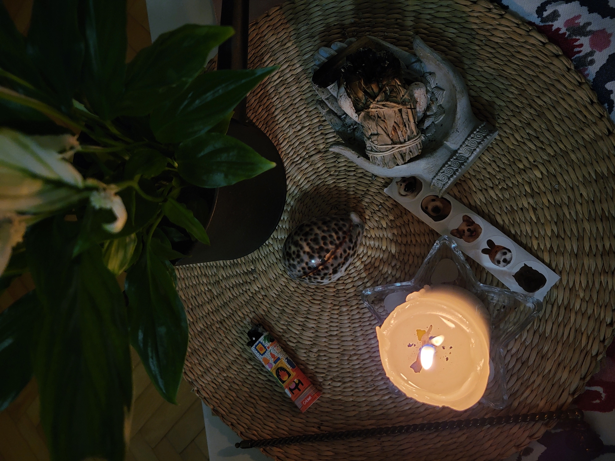 A high-angle shot displays a scene indoors. A potted plant with large green leaves occupies the left of the image, set on a dark table. A round, woven straw mat fills the right half. On the mat, an array of objects are arranged: a decorative hand sculpture holding what appears to be sage, a patterned shell, a lighter with colorful illustrations, and a lit candle in a clear, star-shaped dish. A strip of stickers with dog faces is also present. The edge of a patterned fabric is visible at the bottom right. The lighting is dim, creating shadows and muted tones. The location in the city of Budva, Montenegro is not discernable from this image.