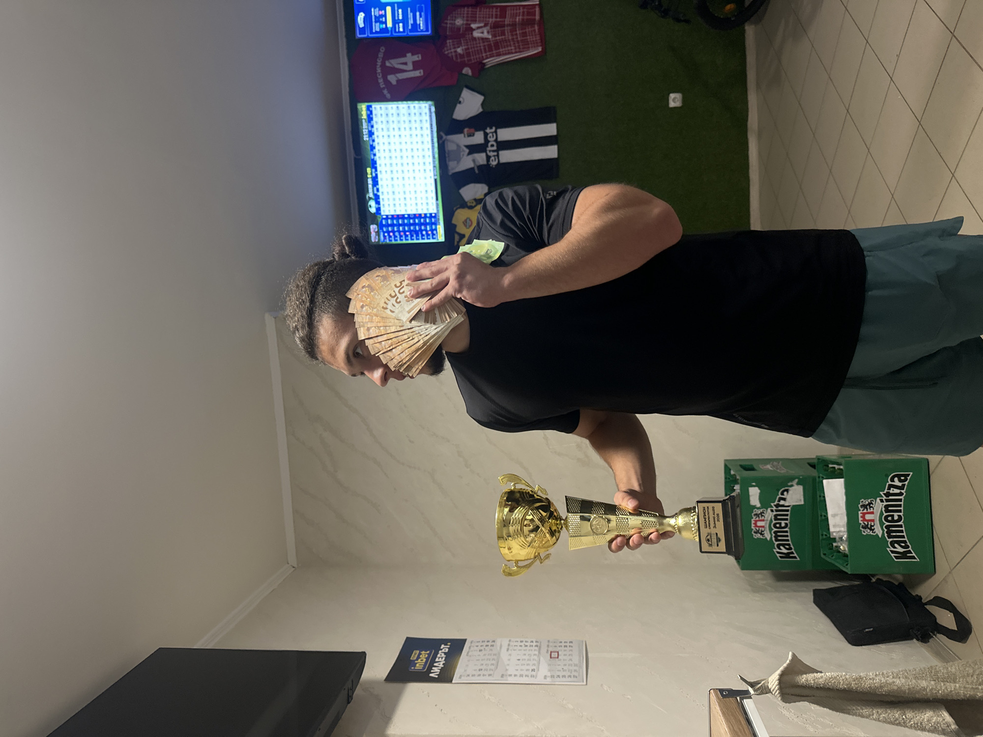 A man is depicted indoors, likely in Ivailo, Bulgaria, holding a large fan of banknotes, appearing to be 50 Euro bills, in his left hand, and a golden trophy in his right hand. He is wearing a black t-shirt and blue-green shorts, with his dark hair tied back. His gaze is directed downwards towards the money and trophy.

The setting suggests a sports-themed room or a space dedicated to sports enthusiasts. The wall behind him is adorned with several sports jerseys, including a red one with "14" and "A1", and a black and white striped one featuring "efbet", a Bulgarian betting company. Two television screens are mounted on the wall, displaying what appear to be scoreboards or betting interfaces. A section of artificial green turf is visible on the wall, further emphasizing the sports theme.

On the floor to the right, two green plastic crates, likely for Kamenitza beer (a Bulgarian brand), are stacked, with the trophy's base resting on them. A black bag is placed next to the crates. On a surface below the trophy, a flyer or calendar shows the text "inbet" and "ЛИДЕРЪТ" (LIDERUT - "The Leader"), another Bulgarian reference. The trophy itself bears inscriptions such as "ШАМПИОН" (CHAMPION), "СПОРТНО ПАНГО" (SPORTS PANGO), and "2023", indicating a sports championship from that year.

The scene conveys a sense of victory or successful achievement, possibly related to sports betting or a competition, given the display of cash and the trophy.