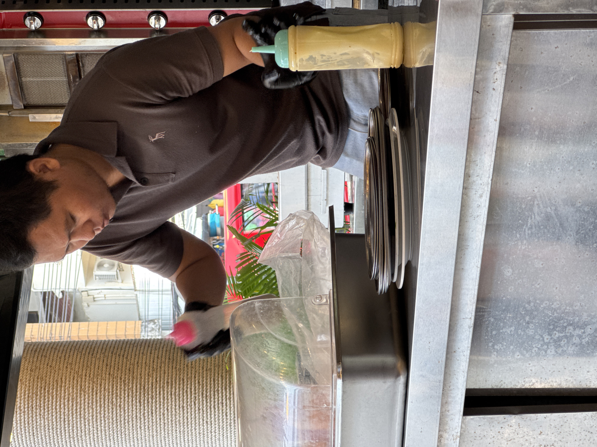In Karon, Thailand, a man wearing black gloves and a brown polo shirt is preparing food. He is holding a pink squeeze bottle in his left hand, with its cap detached. In his right hand, also gloved, he holds a yellow squeeze bottle with a light blue cap. The bottles appear to contain condiments. In front of him is a stainless steel tray with a clear plastic lid, partially covering a transparent container inside. To the right of the tray, a stack of dark plates is visible. The background shows a vertical textured column, possibly a pillar, and foliage, suggesting an outdoor or semi-outdoor setting. The scene appears to be at a food stall or restaurant counter.