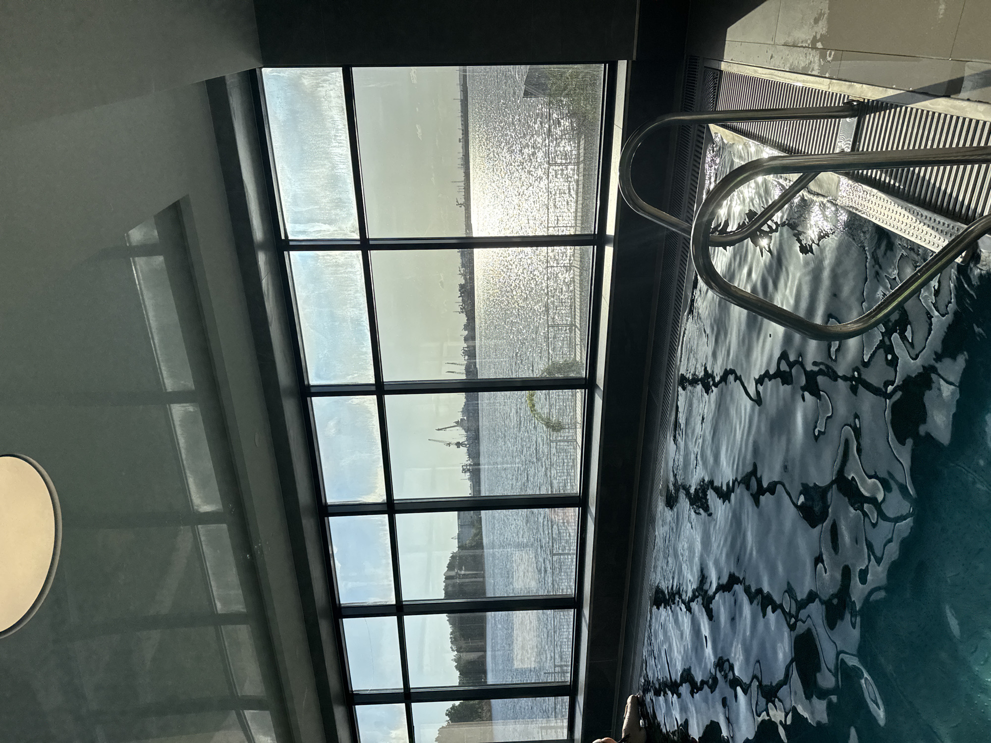 This image shows the interior of a pool area, with a large window displaying a waterfront view. Through the window, a body of water reflects sunlight, and in the distance, cranes can be observed, which suggest a port or industrial area. The location is Chaikovsky, Russia. The pool area itself is tiled with dark grey and the ladder used to enter the pool is in the right corner. The view is from a high level, as the pool, the window, and the body of water are visible and the view is downward. There is an off-white light in the upper left corner. The reflections of the window can be seen on the ceiling.