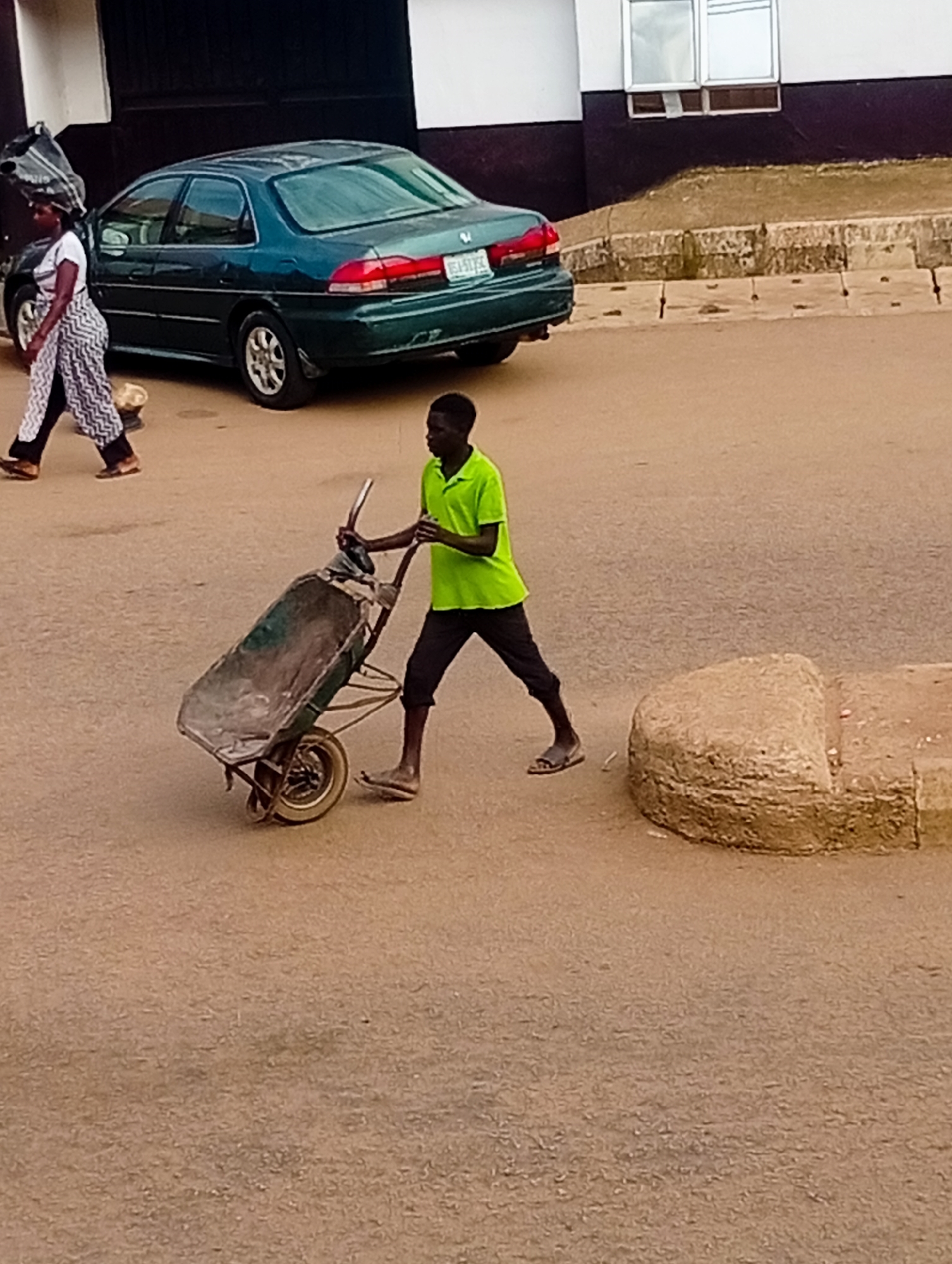 The image depicts a street scene in Bukuru, Nigeria. In the foreground, a young man is pushing a wheelbarrow. He is wearing a bright green t-shirt and dark trousers. The wheelbarrow appears to be old and is loaded with some material, possibly soil or debris, covered with a grey tarp. To the left, a woman is walking, carrying a large bag on her head. She is wearing a white t-shirt and a patterned skirt. Behind the woman, a dark green sedan car is parked. The car has a license plate that appears to read "BEA 51 PC". The background shows a building with windows and a concrete structure bordering the road. The ground is paved and appears to be dusty. The overall scene suggests everyday life and activity in the city.