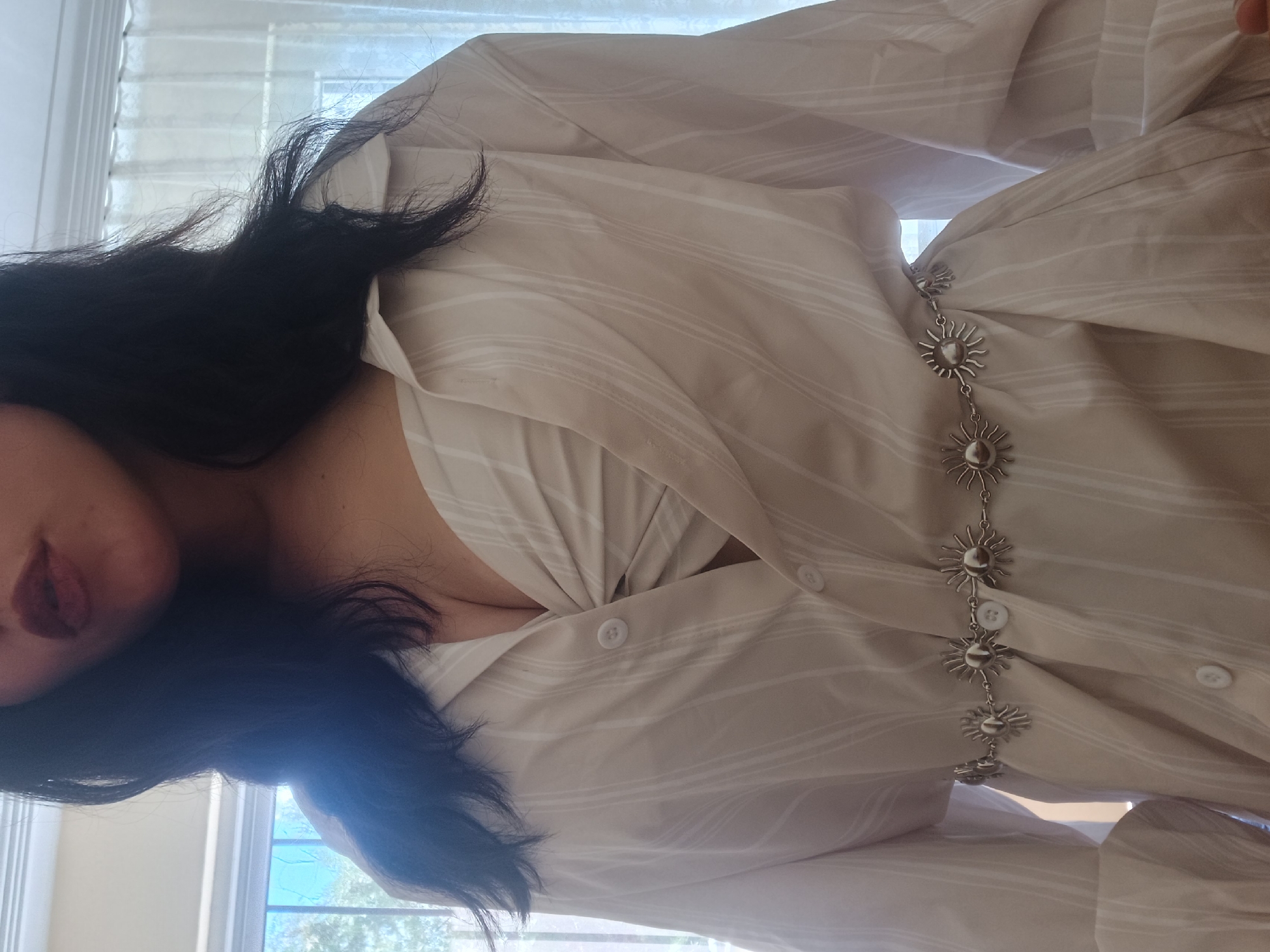 A woman with dark hair is wearing a light beige striped shirt with a silver-colored chain featuring sun-shaped pendants hanging down the center of the shirt. The background shows a window with light coming through. The location is not discernible as Sofia, Bulgaria.