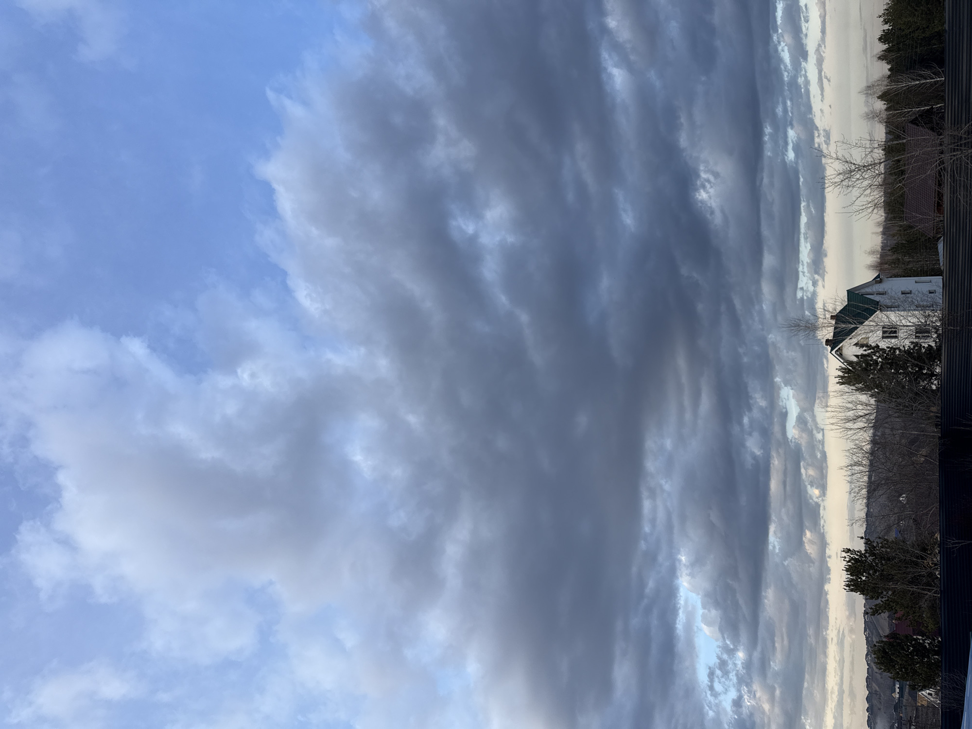The image shows a view from Бугачёво, Russia, looking towards a cloudy sky. The clouds are a mix of light blue and dark gray, with some areas showing breaks in the cloud cover. Below the clouds, there are several houses and trees. The houses are a mix of colors, with one house being white with a green roof. The trees are mostly bare, indicating that it is likely winter or early spring.