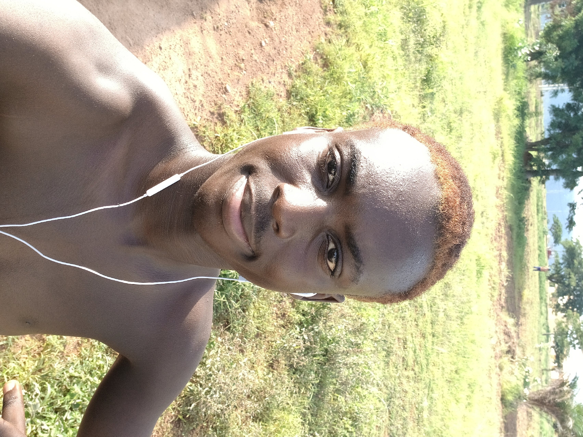 A medium shot selfie captures a dark-skinned young male with cropped, reddish-brown hair. He is shirtless, wearing white earbuds, and looking directly at the camera with a faint smile. His skin is shiny with perspiration. He is positioned in what appears to be an outdoor, grassy area. The background consists of a field of dry grass and scattered trees, with a hint of a body of water in the distance. The overall lighting suggests it is daytime.