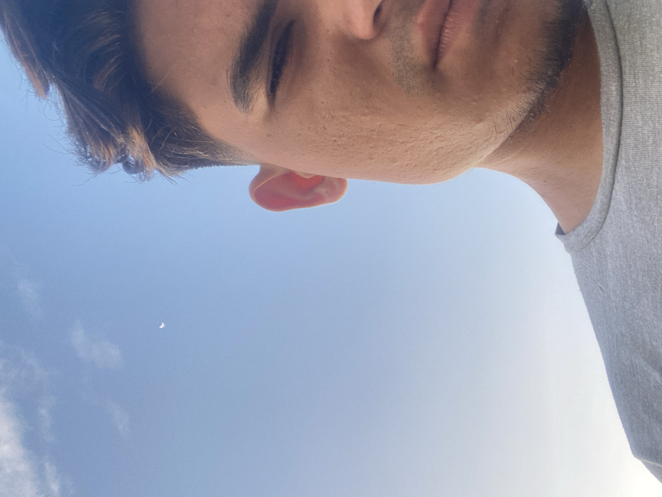 A close-up angled selfie of a man's face, filling the top portion of the image. He has dark hair, light skin, and is partially turned to the side with his eyes closed. A gray t-shirt appears on the right edge. Below the man's face is a bright blue sky with a small crescent moon visible on the left side. Some faint, scattered clouds are visible in the lower half of the sky. The overall composition is upward-facing.