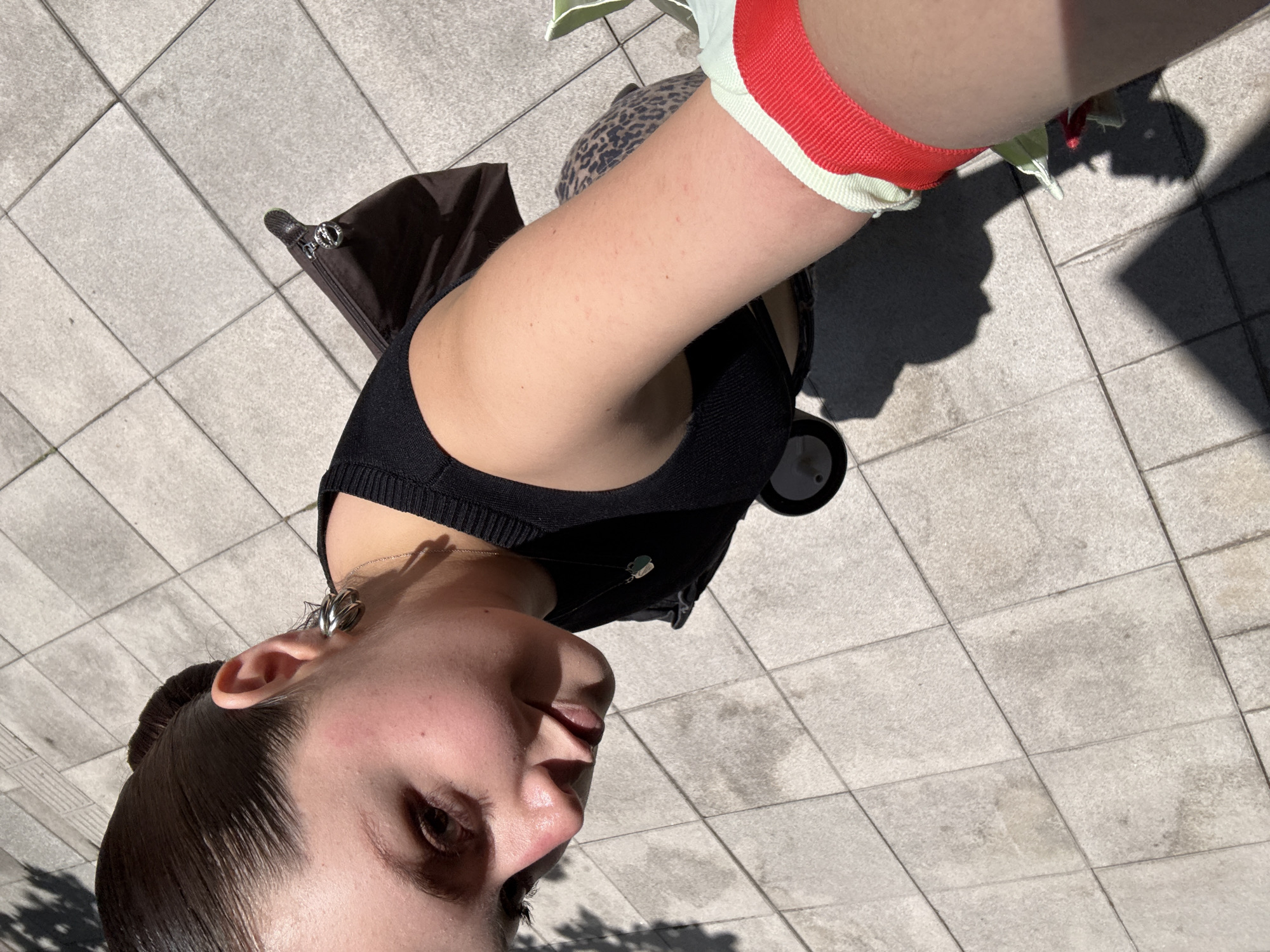 An overhead, tilted shot captures a person outdoors in Mexico City, Mexico, under bright sunlight, evidenced by strong, defined shadows on the ground. The perspective is from above and to the person's right, showing their left profile and right arm prominently.
The subject is a person with dark hair slicked back, wearing a black top or bra-like garment. Their left ear features a distinctive spiral-shaped silver earring, and a delicate silver chain necklace with a small, light green or blue pendant is visible around their neck. The right arm is bare, and around its bicep is a red band with a white stripe. A textured leopard print fabric is partially visible beneath the right arm, possibly belonging to an accessory or another part of their attire. Behind the person's left shoulder, a dark brown bag or backpack with a silver zipper pull is present.
The setting is an urban outdoor space with light grey square paving tiles covering the ground. The strong shadows suggest a sunny day, likely during the mid-morning or early afternoon. The person's expression is neutral, looking towards the right side of the frame. There is no visible text in the image.