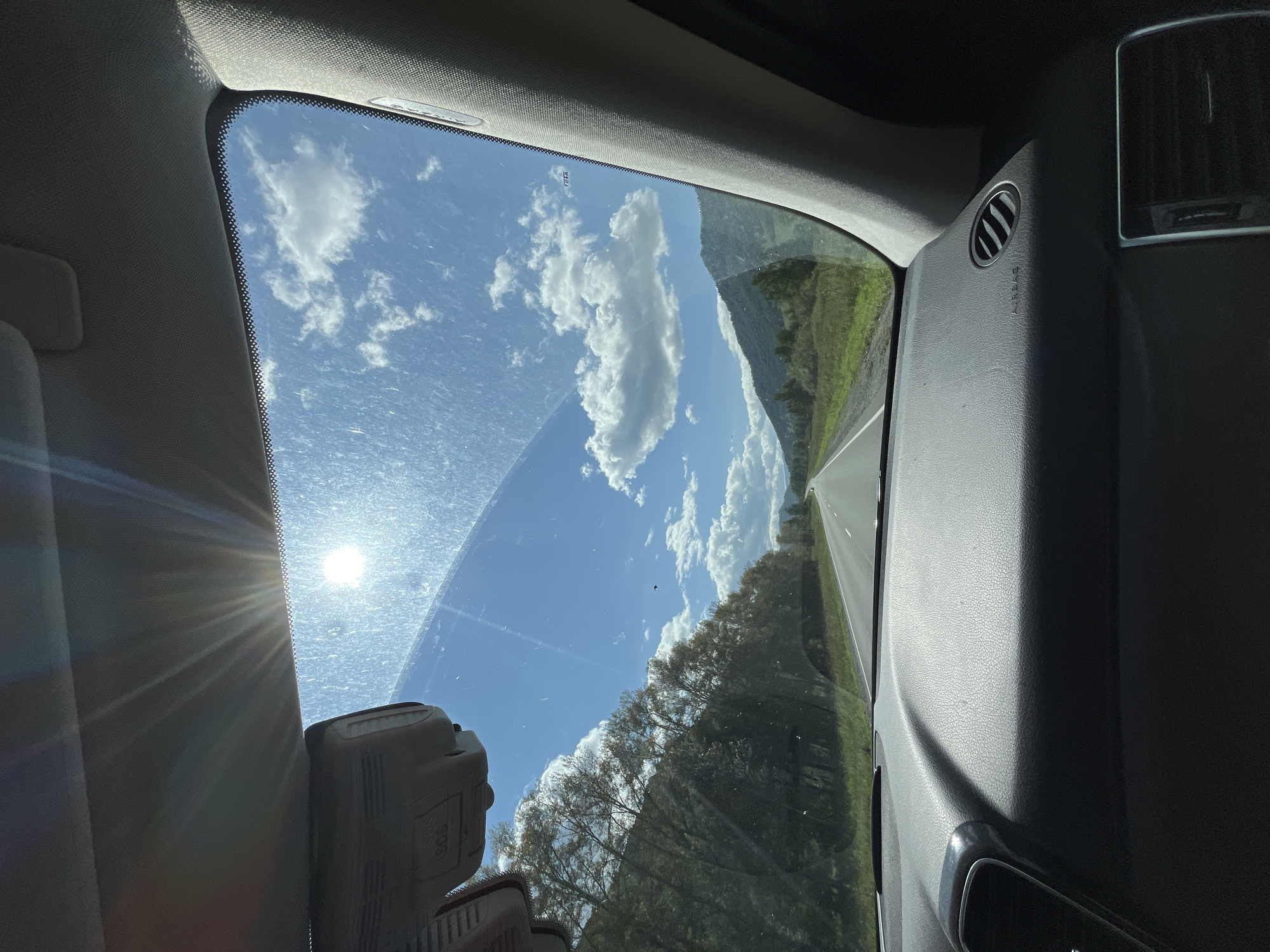 The image, taken from the interior of a vehicle, captures a view through the windshield. The foreground shows the dark, textured surface of a car dashboard with an air vent and an airbag label visible. The windshield itself is slightly dusty, with streaks and reflections. Beyond the windshield, a bright sun with visible rays shines in a blue sky dotted with fluffy white clouds. A road stretches into the distance, flanked by green grassy fields and trees on the left, and rolling green hills and mountains on the right. The scene suggests a journey through a scenic, possibly rural, landscape under a clear, sunny sky. The provided crops offer closer views of the sun, the sky, the interior of the car, and the roadside scenery. The location is stated as Myiuta, Russia.