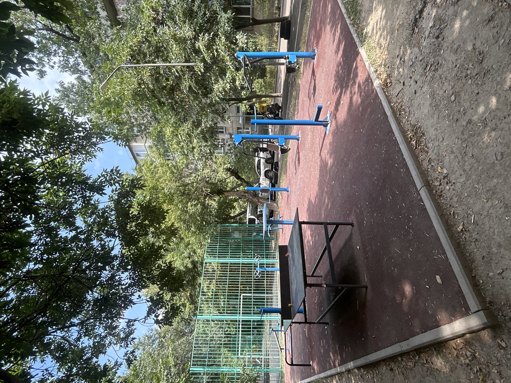 An outdoor sports court in Almaty, Kazakhstan, features a ping pong table with a dark surface and metal frame. Adjacent to the table is a green metal fence, likely for a sports field, with trees and buildings visible in the background. The court itself has a red, rubberized surface. Several blue exercise machines, including pull-up bars and leg presses, are arranged on the court. A street lamp stands on the right side of the frame. The ground surrounding the court is dirt and scattered with leaves. The scene appears to be a public recreation area, possibly a courtyard in a residential complex, on a sunny day.