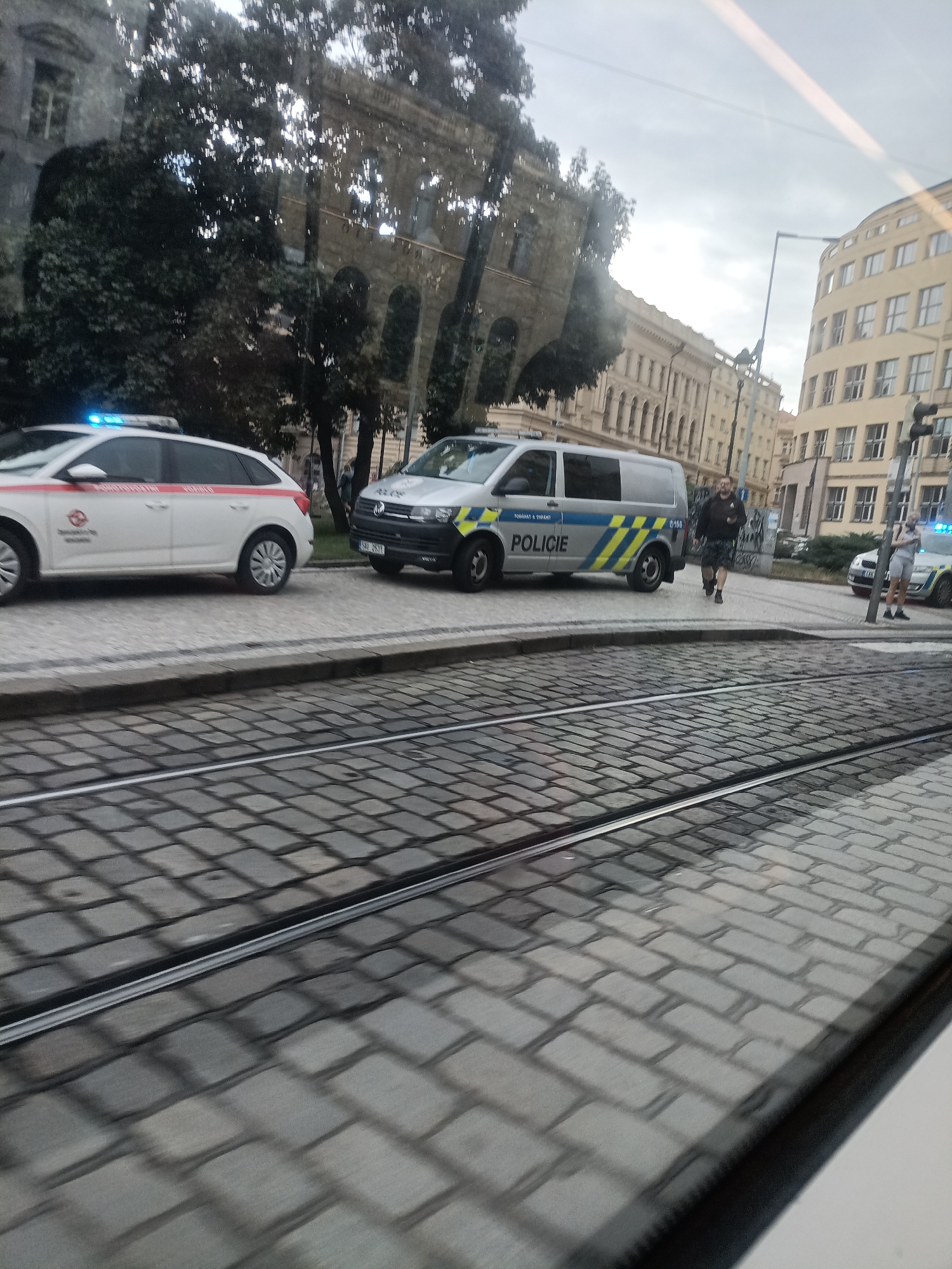 In Prague, Czechia, two police vehicles are present on a cobblestone street. One is a white SUV with red and white markings and a flashing blue light on top. The other is a silver van with yellow and blue checkered stripes and the word "POLICIE" in blue lettering. A man in a dark jacket and shorts is walking away from the van, and a woman in a light-colored outfit is standing near the SUV. Buildings with architectural details are visible in the background. Tram tracks are in the foreground, indicating the location is likely a street with public transportation. The sky is overcast.