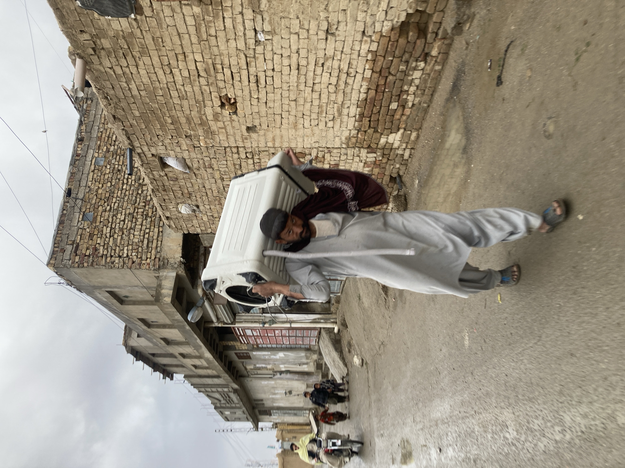 The image shows two men carrying a white appliance, likely a washing machine, through a narrow alley in what appears to be a densely populated area of Quetta City Tehsil, Pakistan. The alley is flanked by weathered brick buildings. One man is wearing a light-colored traditional shalwar kameez and a dark cap, bearing the weight of the appliance on his shoulder. The other man is wearing a maroon robe and assisting with carrying the load. A group of children is visible in the distance, along with a motorcyclist. The road surface is paved and appears wet. The sky is overcast, indicating a potentially damp or rainy day.