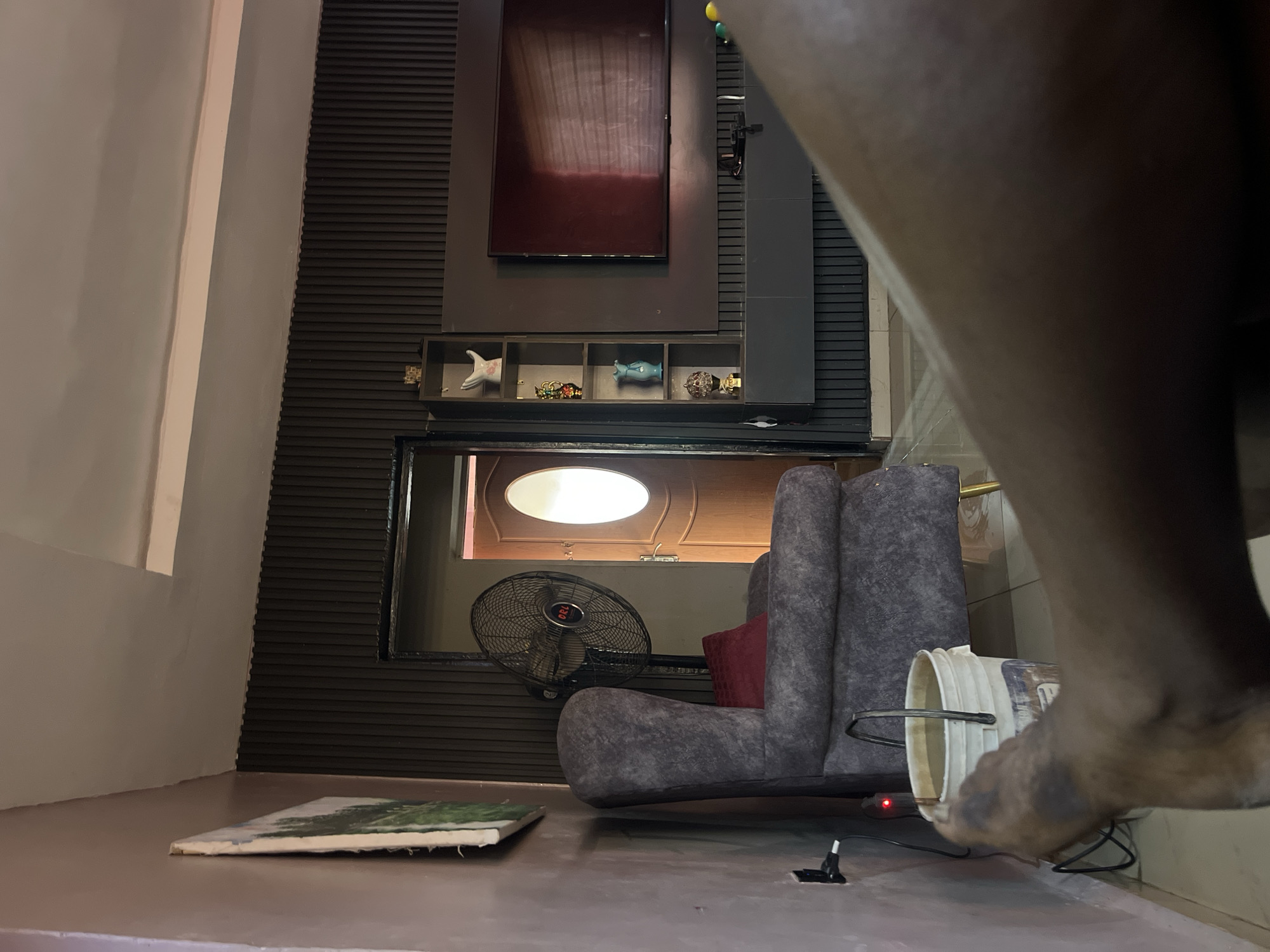 This is a medium shot, indoor photograph taken in a living room. The room has dark, textured walls and a pale ceiling. On the left wall hangs a framed painting of a landscape. In the center of the room, a tall, grey upholstered armchair with a dark red cushion sits in front of a large, black oscillating fan. To the right of the fan, a doorway leads into another room, revealing a wooden door with an oval window. A tall, dark shelf unit with several decorative items is mounted on the wall to the right of the doorway. A large, flat-screen television is mounted on the wall above a dark cabinet below the shelves. In the foreground, a person's bare leg and foot are visible, with the foot appearing dirty. To the left of the foot is a white bucket filled with water. A black electrical cord runs from an outlet on the wall to the fan, with a small red indicator light visible on the cord. The overall impression is of a lived-in space.