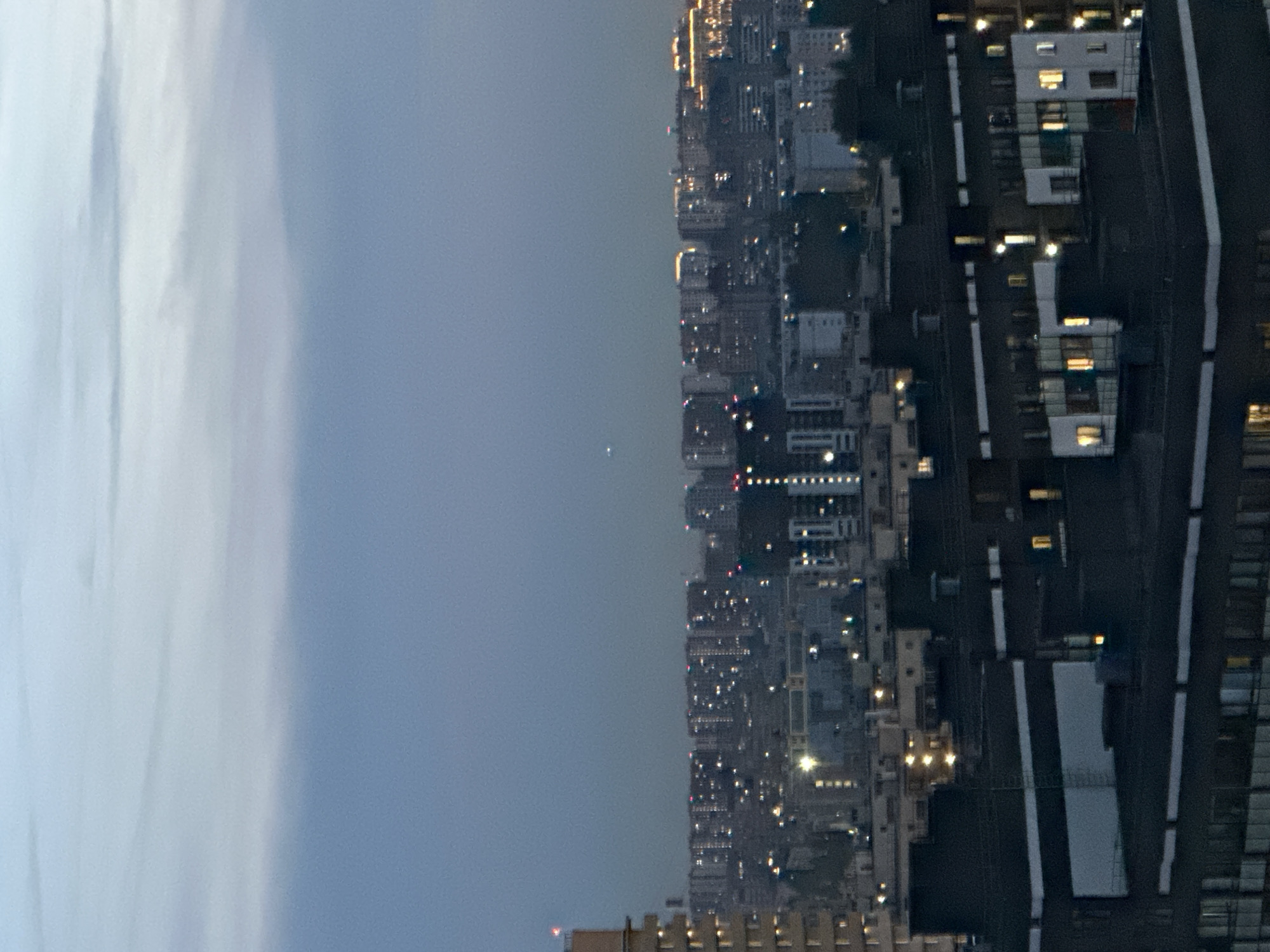 The image depicts a cityscape at dusk or dawn, with buildings of varying heights and architectural styles. The sky is a gradient of pale blue and gray, with some faint cloud formations visible. In the foreground, dark-colored apartment buildings with illuminated windows suggest human habitation. Further in the distance, a dense urban sprawl is characterized by rows of illuminated buildings, some with warm yellow lights, indicating residential or commercial activity. A faint, bright point of light, possibly a star or planet, is visible in the sky above the distant city. The overall impression is of a large, active city, likely at a time when artificial lights are beginning to dominate the natural light. The scene is static, with no visible people or active events. The location is Feklistovo, Russia.