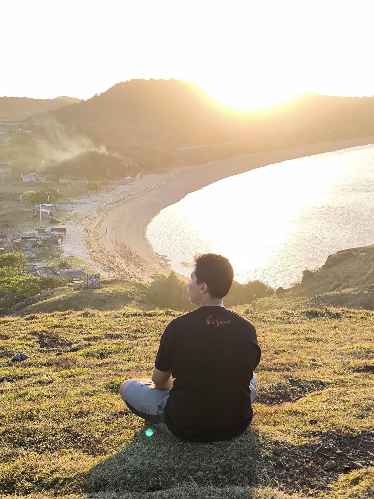 A man sits cross-legged on a grassy hill overlooking a bay in Kuta, Indonesia. The bay has a sandy beach and calm, reflective water. A small village sits at the edge of the beach, partially obscured by light smoke or haze. The sun is setting over a distant hill, casting a golden glow over the entire scene. The man is wearing a black t-shirt and gray shorts. The grass on the hill is dry and yellowed.