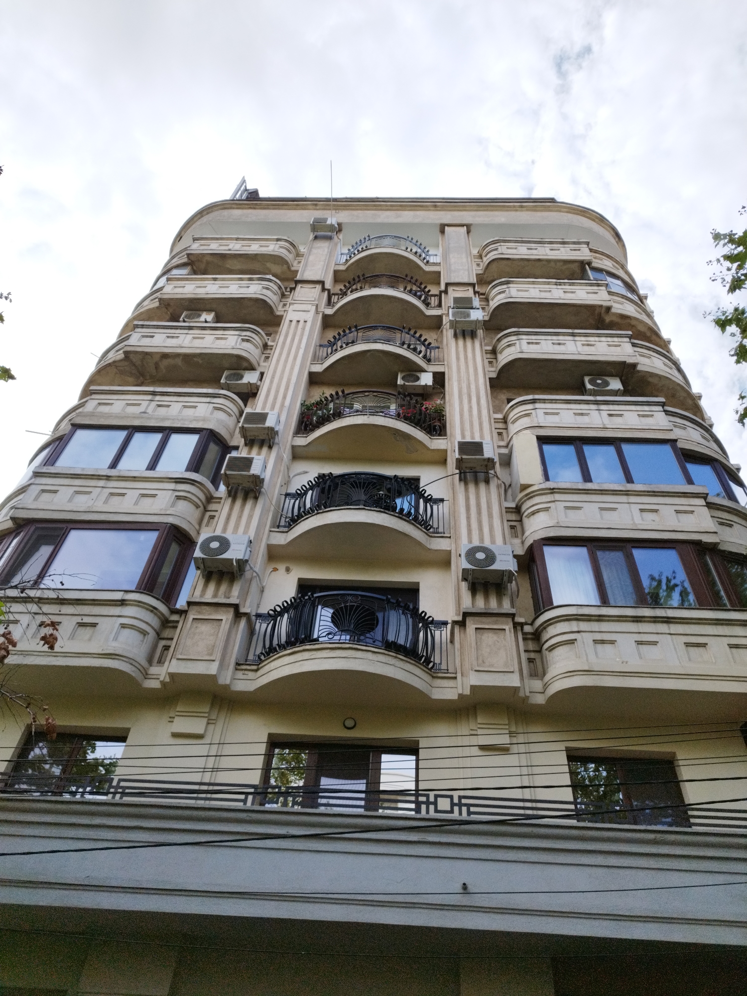 The image shows the exterior of a cream-colored, multi-story building with a curved facade, located in Bucharest, Romania. The building features numerous balconies with black wrought-iron railings, and several windows with dark frames. Air conditioning units are mounted on the exterior walls between floors. The sky above is overcast with clouds. The photograph is taken from a low angle, looking upwards at the building.