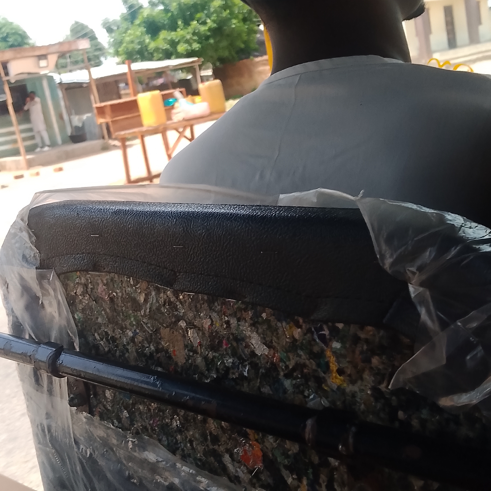 The image is a close-up of the back of a seat, possibly in a vehicle, with its internal stuffing exposed and wrapped in plastic. In the background, there are stalls with wooden shelves and yellow containers, suggesting a market or roadside vendor setting. A person in a light-colored garment is visible in the distance, standing near one of the stalls. The scene is outdoors, with trees and structures indicating an urban or semi-urban environment. The overall impression is one of everyday life in a community, possibly a market in Jalingo, Nigeria, given the context provided. The focus is on the damaged seat, with the background offering a glimpse of the surroundings.