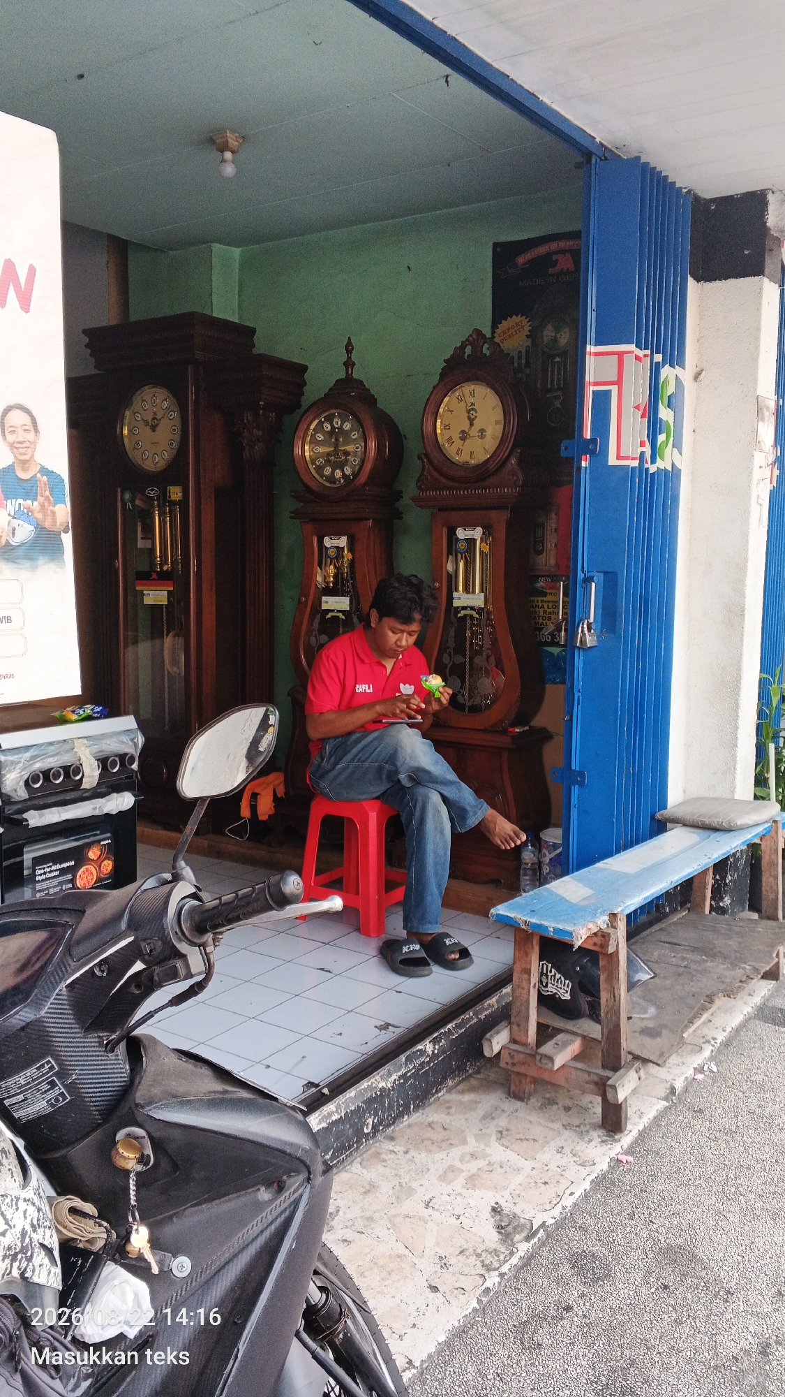 A man in a red polo shirt and blue jeans is sitting on a red stool in front of a shop, eating something green. He is looking down at his phone. Behind him are three large grandfather clocks. The shop appears to be in Malang, Indonesia, judging by the timestamp on the image. To the left of the man is a motorcycle, and to the right is a wooden bench with a blue seat. The shop has a blue metal gate that is partially closed. The ground is tiled. The wall of the shop is painted green.