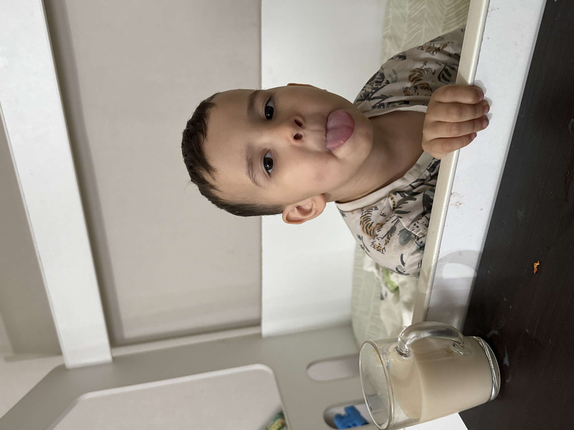 A child is sticking their tongue out at the camera. The child is wearing a patterned shirt and grasping a white railing. A clear glass mug containing a light-colored liquid sits on a dark surface. The scene appears to be indoors, possibly within a home. Location information (Chaikovsky, Russia) cannot be verified from visual data.