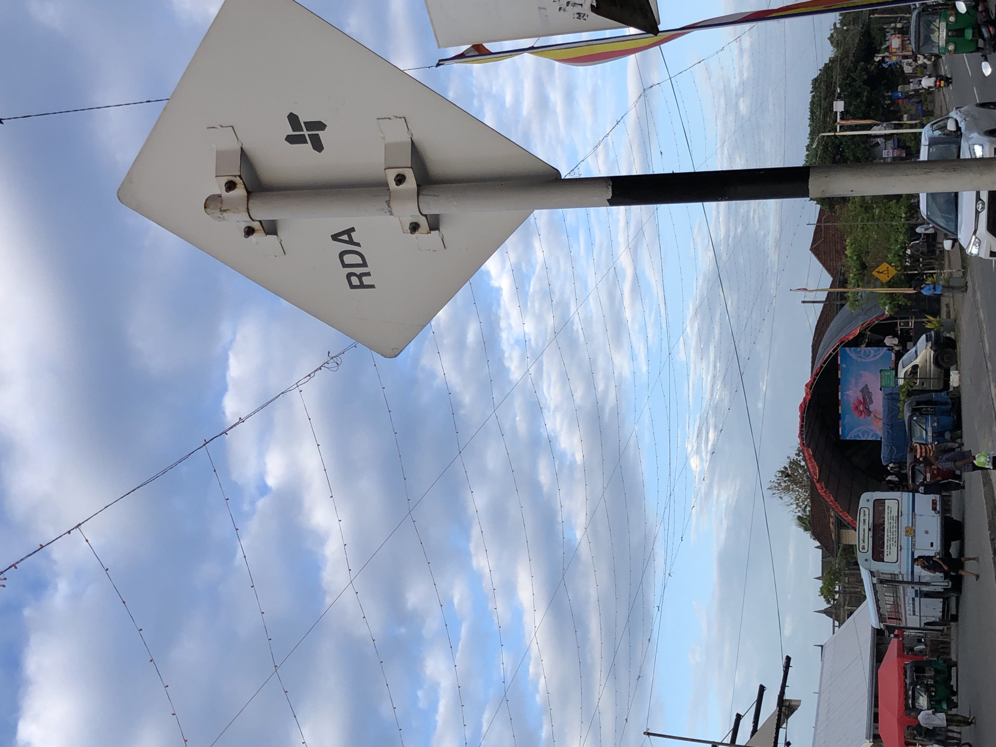 The image shows a street scene in Habaraduwa, Sri Lanka, viewed from a low angle looking upwards towards the sky and the side of a street. A white sign with black text that reads "RDA" is prominently displayed. Above the sign, strings of small lights are strung across the sky, creating a decorative effect. In the background, there is a street with various vehicles, including tuk-tuks and cars, and some buildings. The sky is partly cloudy with a mix of blue sky and white clouds. The lighting suggests it is daytime, possibly late afternoon or early evening. There are people visible on the street and sidewalks. The overall impression is a vibrant street scene in a tropical location.