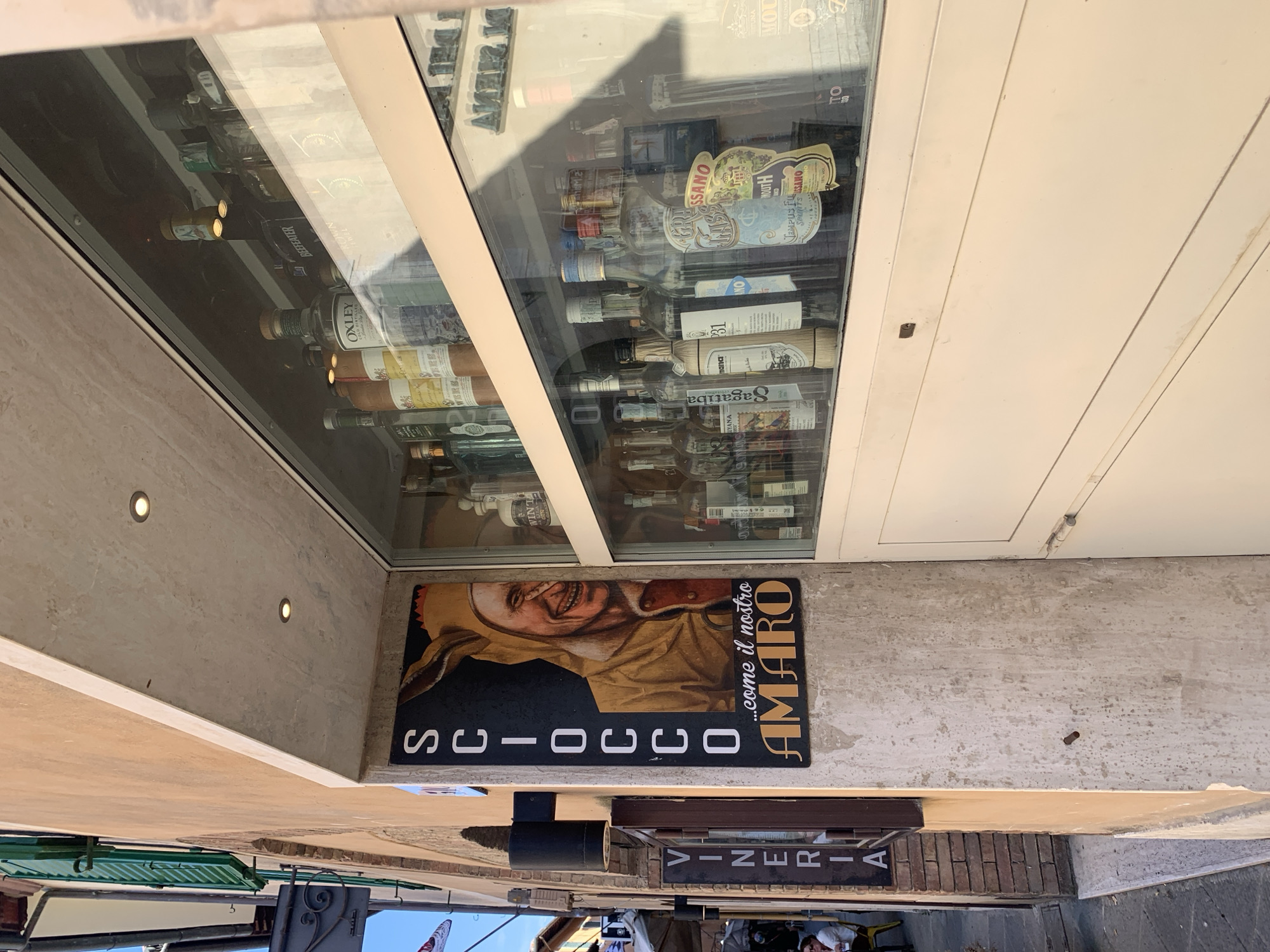 The image shows the exterior of a shop in San Quirico d'Orcia, Italy. A tall, vertical sign with the words "SCIOCCo ...come il nostro AMARO" is mounted on the wall, featuring a painted image of a person in a jester-like costume with a wide smile. Next to this sign, a large display window showcases an assortment of bottles, presumably alcoholic beverages, arranged on shelves. One of the bottles in the window has a label that reads "GRASSO". A narrow, darker sign with the word "VINERIA" is also visible on the wall below the larger sign. The shop facade is a warm, peachy color, and the street in front of it is paved with cobblestones. In the background, a person is seated at a table outside, and architectural elements of the town are visible.