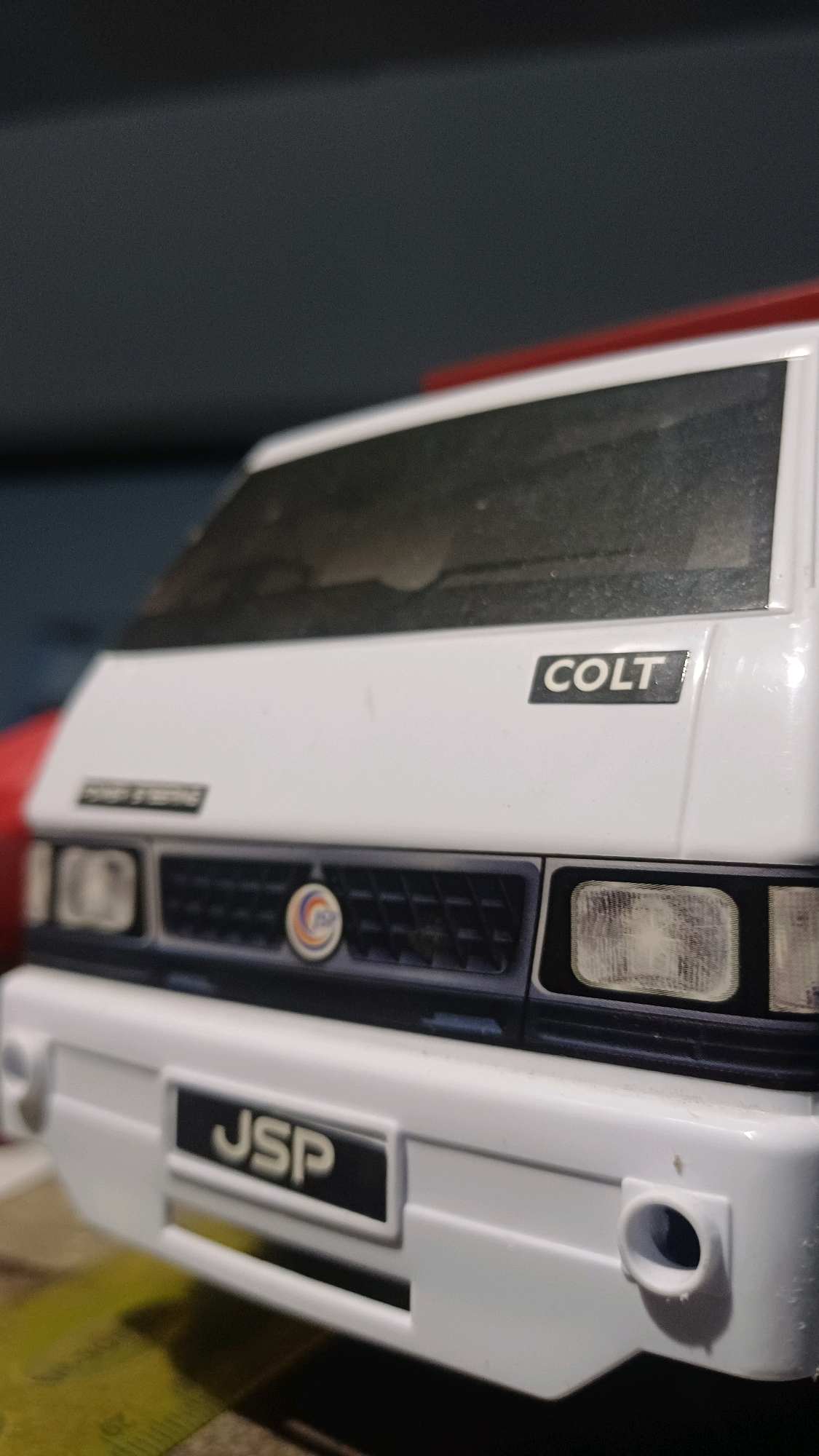 A close-up, low-angle shot focuses on the front of a white model van, possibly a Mitsubishi Colt. The van features a black grille with a circular emblem in the center, and two headlights. The word "COLT" is printed in black on a rectangular badge above the grille. Below the grille, the letters "JSP" are displayed on another black badge. The background is blurred, showing a dark surface and a hint of red. The scene is not located in the city of Wonokerso, Indonesia.