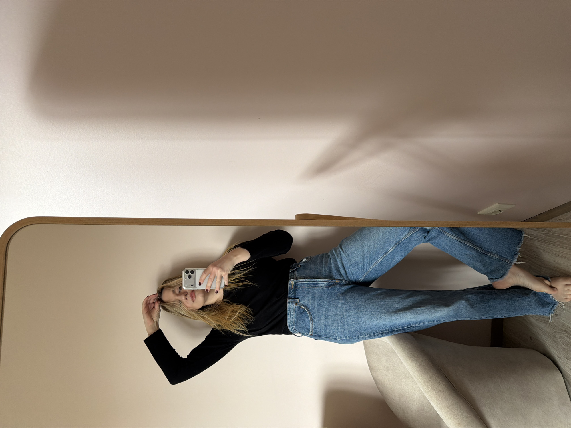 A woman is reflected in a mirror, holding a smartphone, presumably taking a selfie. She is wearing a black long-sleeved shirt and blue jeans. The mirror has a wooden frame. Part of a beige chair is visible at the bottom right corner. The background behind the mirror is a light beige wall. The exact location within Dreiliņi, Latvia, cannot be determined from the image.