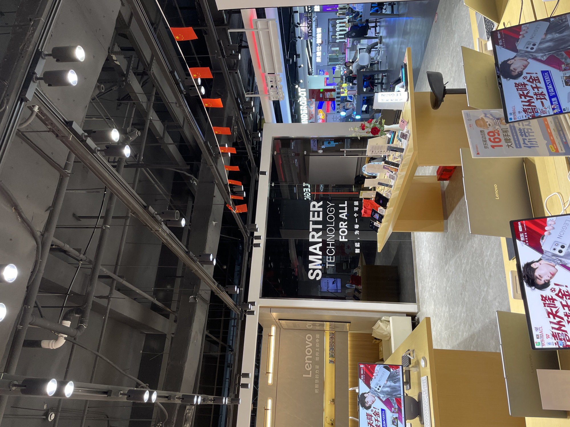 The image shows the interior of a modern electronics store, likely a Lenovo store, located in Liangjiang, China. The store is brightly lit with overhead track lighting and features a clean, organized display of electronic products, including laptops and possibly phones, arranged on tables. A large sign on the back wall reads "SMARTER TECHNOLOGY FOR ALL" in English, with Chinese text below. Chinese flags are strung across the ceiling. In the foreground, several laptops and displays are visible. In the background, customers can be seen browsing and interacting with the products. The overall impression is of a well-maintained retail environment showcasing technology.