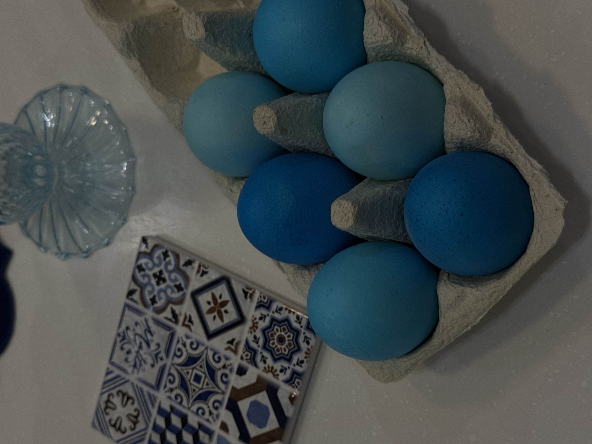 The image depicts a still life arrangement on a white surface. A cardboard egg carton contains six blue-colored eggs. A small, blue-tinted glass vase is positioned to the left of the carton. A square tile, patterned with various blue and white geometric designs, rests below the vase and to the left of the carton. The setting appears to be indoors. There is no evidence of the location being Khimki, Russia.