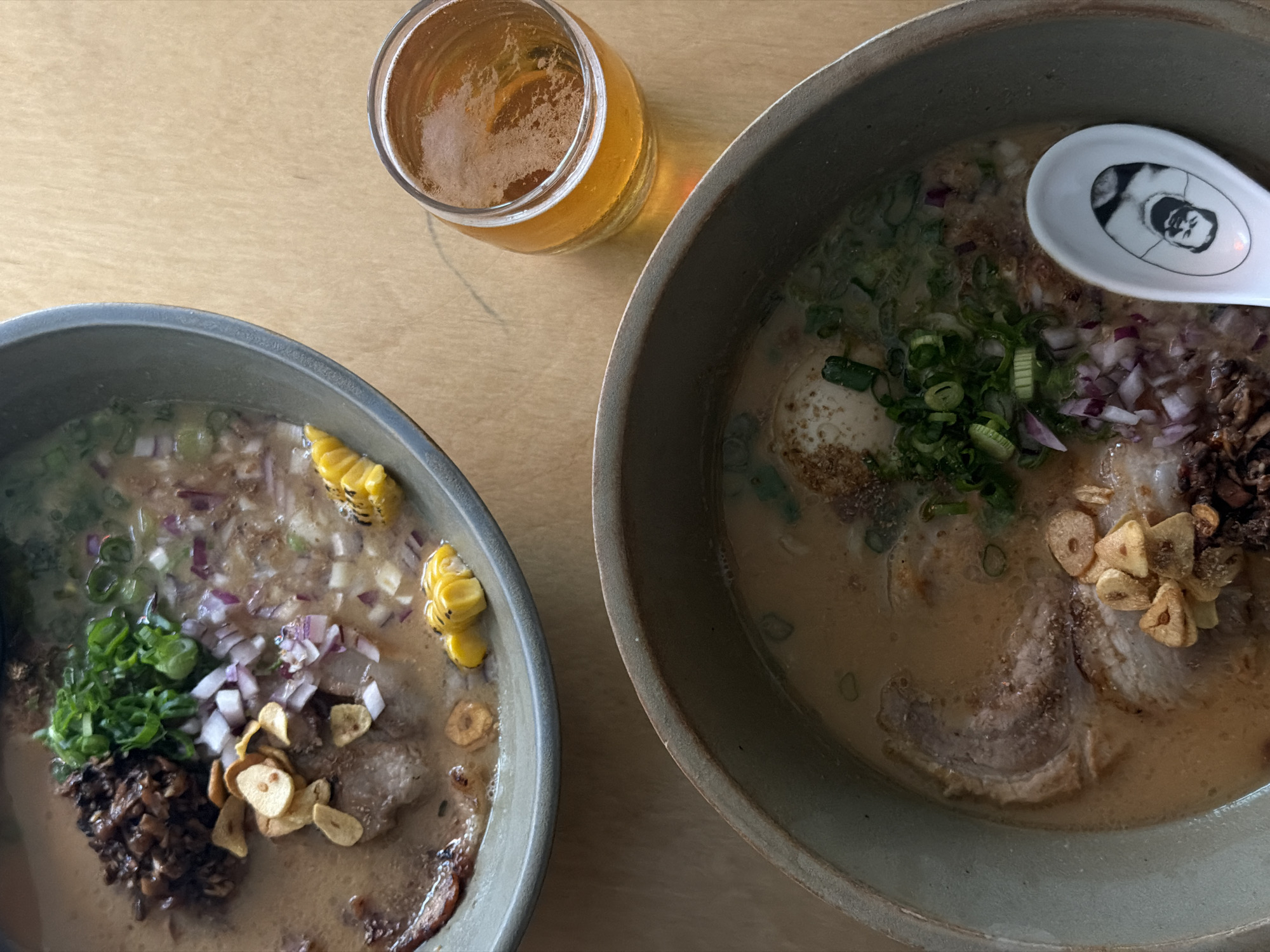 Two bowls of ramen and a glass of beer are on a wooden table. The ramen bowls are stoneware in a matte gray-green color. The ramen contains visible components such as sliced pork, green onions, diced red onion, fried garlic chips, and corn. One bowl has a white ceramic spoon with a picture on it. The beer is in a clear glass with visible condensation on the outside. There are no people visible in the scene. The lighting is soft and diffused. It can't be determined that the location is in the city of Belgrade, Serbia.