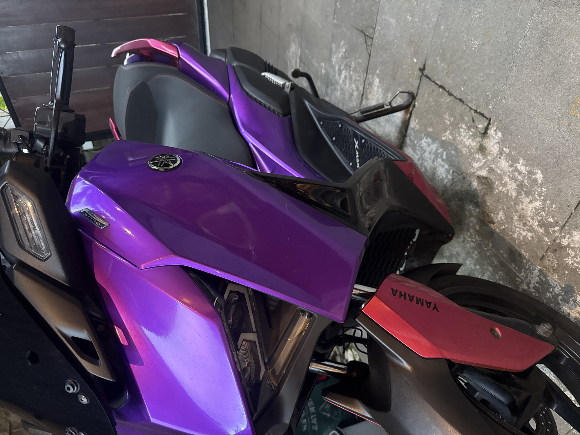 A close-up shows two Yamaha motorcycles parked side by side near a concrete wall in Ubud District, Indonesia. The motorcycle in the foreground is predominantly purple, featuring a Yamaha logo on the side. A red fender with "YAMAHA" printed on it is visible near the rear tire. The other motorcycle, mostly out of focus, shares the same purple color scheme. The scene appears to be in an urban or semi-urban setting, given the presence of concrete structures.