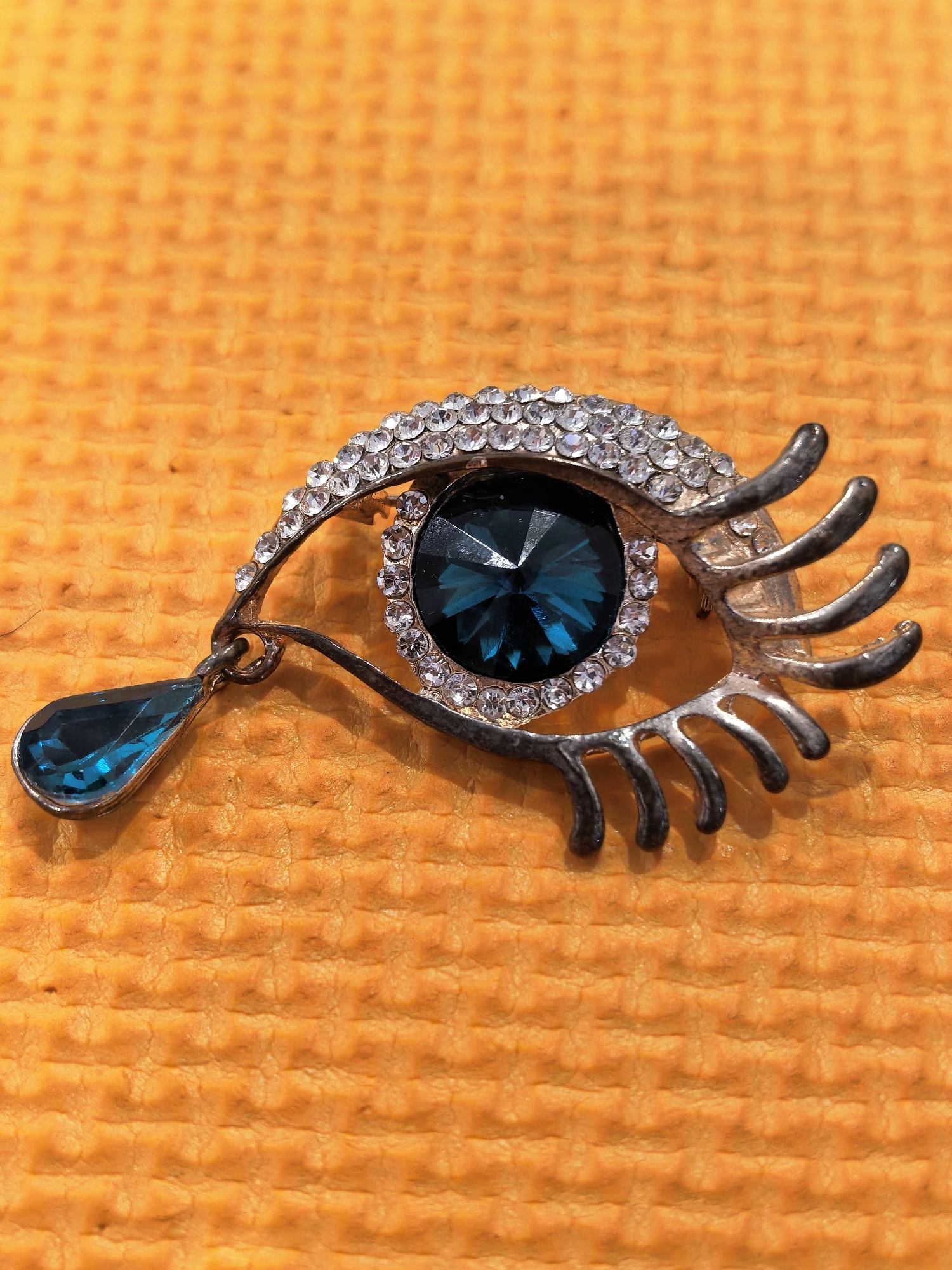 A brooch shaped like an eye is displayed on an orange, textured surface. The brooch features a dark blue, faceted gem as the pupil, surrounded by a layer of small, clear rhinestones that form the eyelid. The metallic structure of the brooch is silver-toned, designed to resemble an open eye with eyelashes. A teardrop-shaped blue gem hangs from the outer corner of the eye. The orange surface beneath the brooch has a uniform, dimpled pattern.