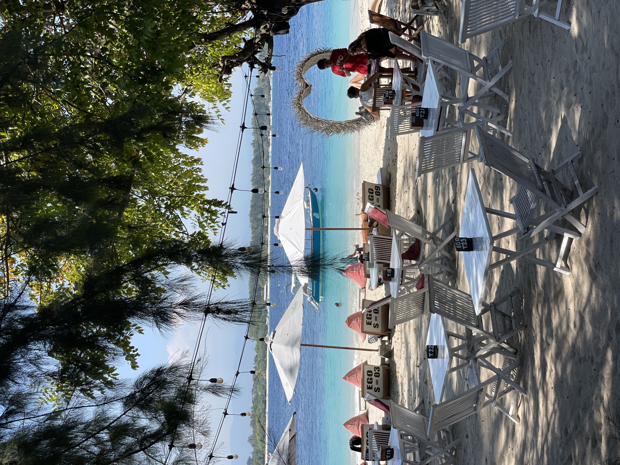 An outdoor cafe on a sandy beach in Unknown, Indonesia, features a tranquil scene with the ocean in the background. Several white tables and chairs are arranged on the sand, some set with table numbers. A heart-shaped decoration made of twigs stands near the water's edge. A group of people are seated at a table, interacting with each other. A boat is anchored in the clear blue water, and umbrellas provide shade. Trees with lush green foliage overhang the cafe area, casting shadows on the sand. String lights are hung overhead, adding to the ambiance. The overall atmosphere is relaxed and inviting, suggesting a perfect spot for a seaside dining experience.