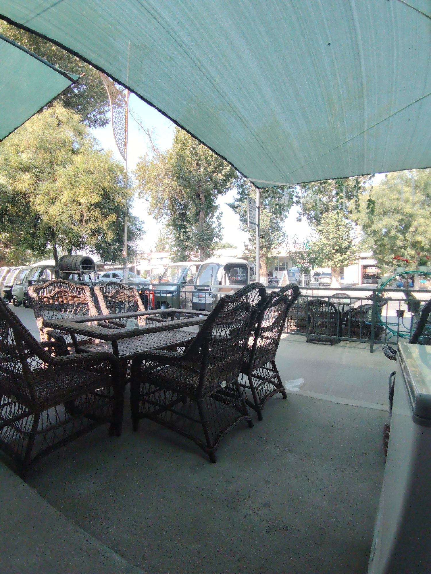 The image depicts an outdoor seating area in Ibrat, Uzbekistan. In the foreground, a table and several wicker chairs are arranged on a concrete floor. The chairs and table are made of a dark, woven material, suggesting they are intended for outdoor use. Behind the seating area, a light green fabric canopy is stretched overhead, providing shade.
Beyond the immediate seating area, a row of small, light-colored vehicles, possibly auto-rickshaws or tuk-tuks, are parked. These vehicles appear to be a common mode of transportation in the area. In the background, trees with lush green foliage line a street, and buildings are visible, suggesting an urban or semi-urban environment. The overall scene suggests a commercial establishment, such as a cafe or restaurant, with an outdoor dining or waiting area. The lighting indicates it is daytime, with sunlight filtering through the trees and canopy. There are no people clearly visible in the image.