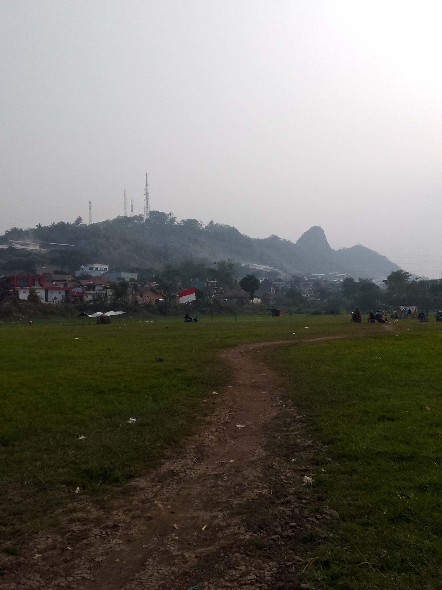 The image depicts a hazy, overcast day in Ciburuy, Indonesia. In the foreground, a dirt path winds through a grassy field dotted with litter. In the middle ground, a collection of buildings, some with red roofs, are nestled at the base of a tree-covered hill. An Indonesian flag is prominently displayed on a pole near the buildings. The background features more houses and buildings spread across the hillside, with several tall communication towers visible. The very distant background is dominated by a prominent, jagged mountain peak shrouded in mist. Several individuals and what appear to be small vehicles are present in the middle ground, some gathered near the buildings, others on the field. The overall impression is one of a semi-urban landscape with elements of rural life.