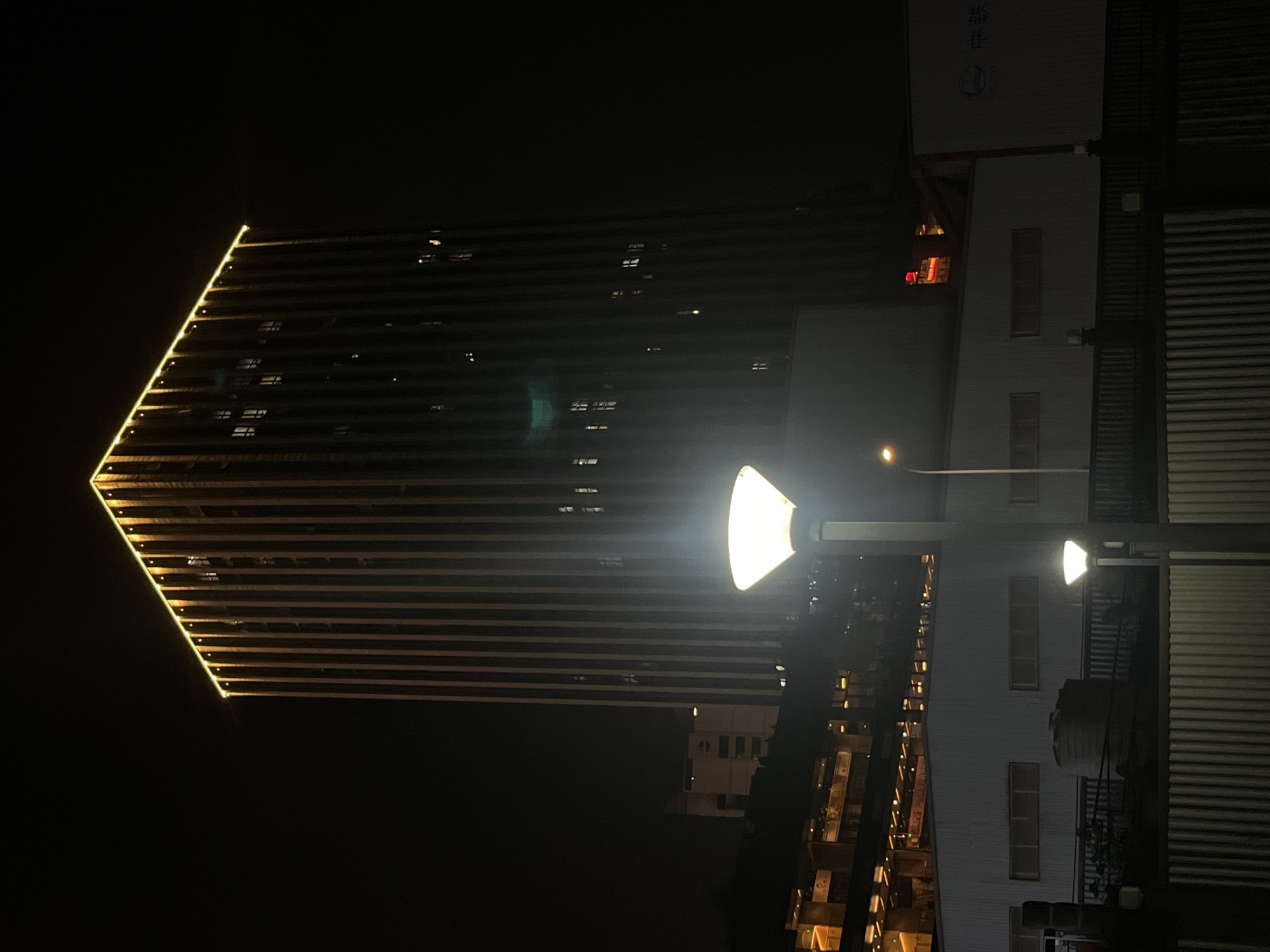 The image captures a nighttime urban scene in Yuzhong District, China, featuring a prominent, tall, rectangular building on the right. This building is characterized by numerous dark vertical panels along its facade and is illuminated by bright yellow LED strip lighting outlining its top edge and descending vertically along its corners. Scattered windows across its dark surface reveal internal lights.
In the foreground, a brightly illuminated street lamp with a white, conical shade stands centrally, emitting significant glare. A smaller, less intense streetlight of similar design is positioned below it. To the left of the main streetlight, a low-rise, light-colored building with several horizontal rectangular windows is visible, partially obscured by a dark metal fence running across the mid-ground. Behind this fence, a cylindrical object, likely a water tank, is present.
Further into the background on the left, additional multi-story buildings with illuminated windows signify other urban structures. On the far right, above the low-rise building, a partially visible red illuminated sign and a white sign bearing the Chinese characters "中铁" (China Railway) are discernible. The sky is dark, indicating night conditions. No individuals or specific activities are depicted in the scene.