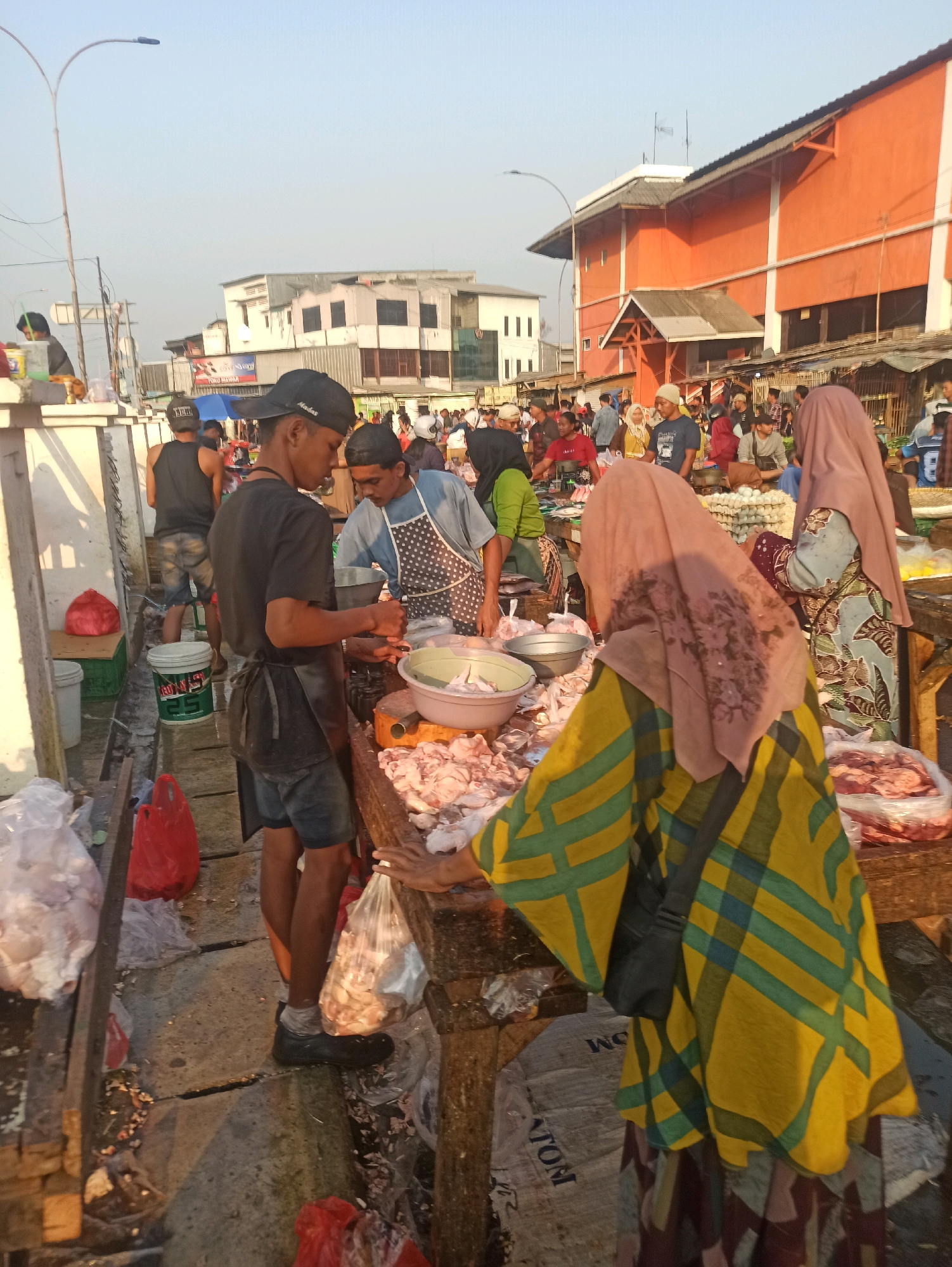 The image depicts a bustling outdoor market scene in Cikarang, Indonesia, likely at a meat stall. Several individuals are actively engaged in transactions and food preparation. In the foreground, a young man wearing a black t-shirt, apron, and a black baseball cap is seen tending to raw meat laid out on a wooden table. He appears to be in the process of packaging or serving the meat. To his right, another person, also wearing an apron, is involved in similar work. A woman in a yellow and green checkered hijab and another woman in a pink hijab are observing or interacting with the vendors. The market is characterized by wooden stalls laden with various cuts of raw meat, some packaged in plastic bags. Other market-goers are visible in the background, suggesting a busy commercial environment. The ground is paved and appears somewhat wet, possibly due to the nature of the market. The overall atmosphere is one of active commerce and daily life in a local Indonesian market.