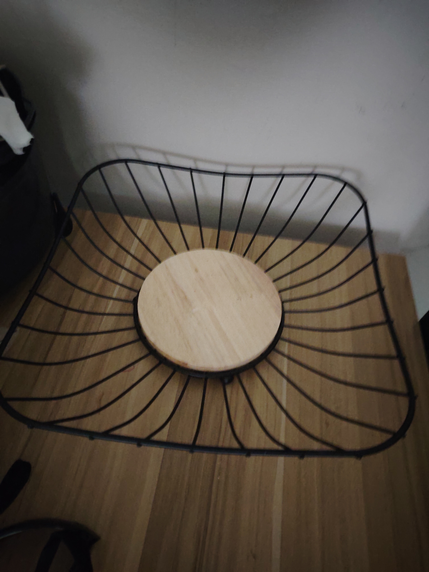 A black metal wire fruit basket with a circular light-colored wooden base is positioned on a light-brown wooden surface. The basket features numerous thin black metal rods radiating from the central wooden disc, curving upwards to form its square-shaped perimeter with rounded corners. The wooden surface displays visible grain patterns. In the upper left background, a dark object, potentially a container, is partially visible, with a crumpled white object resting nearby. The wall behind the objects is plain and light grey. The scene is located in the city of Huntingdonshire, United Kingdom.