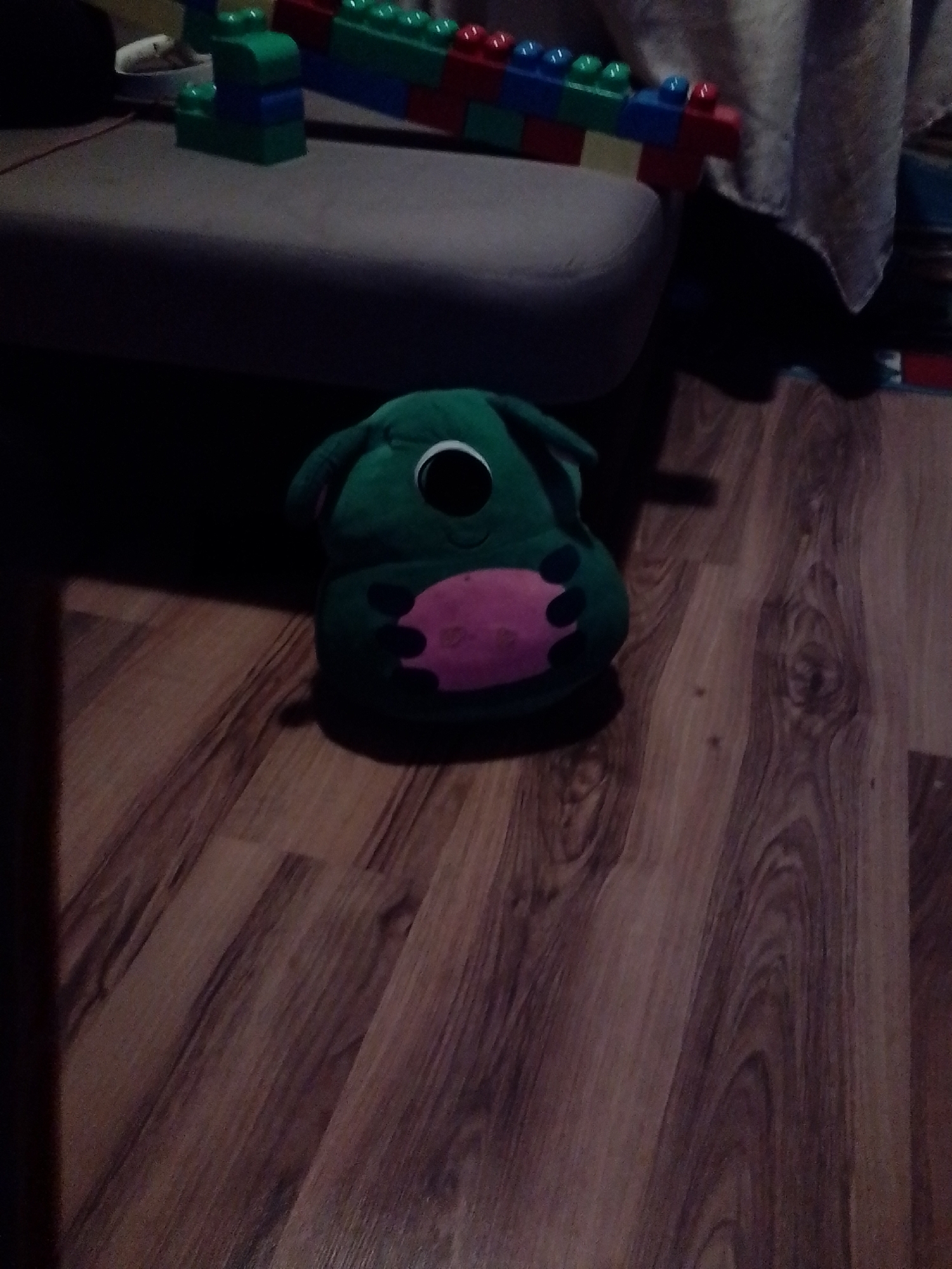 The image shows a green stuffed toy with a large black eye and a pink oval-shaped belly, placed on a wooden floor. In the background, there are building blocks and a gray sofa. There is no indication of the location in Burgas, Bulgaria. The scene appears to be indoors.