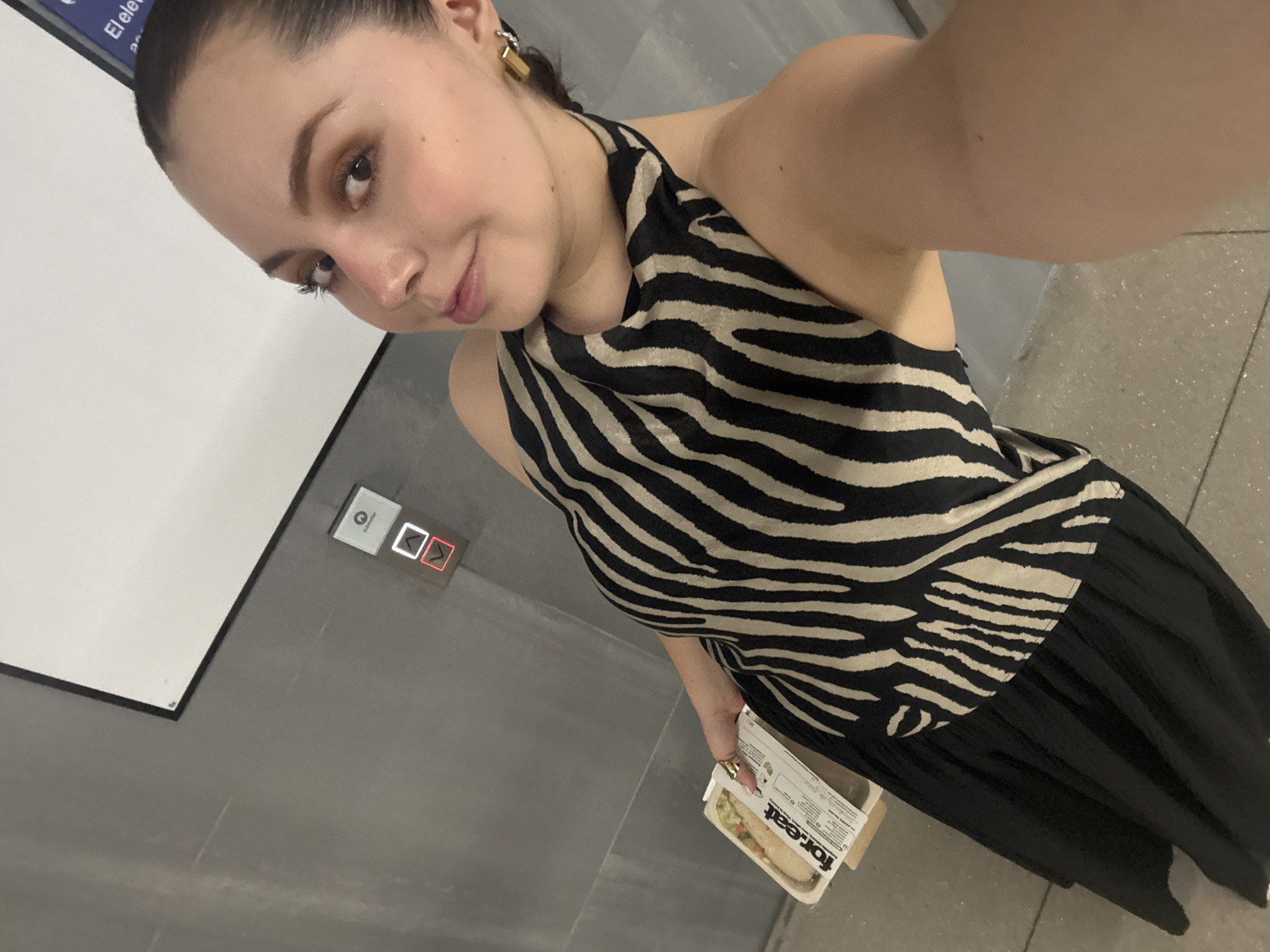 A young woman is taking a selfie in what appears to be a public transportation station. She is wearing a zebra-striped halter top and a black skirt or dress. She has her hair pulled back and is wearing earrings. She is holding a "For.eat" branded container, which appears to hold food. In the background, there is an elevator call button. A sign with the words "El ele..." can be seen partially in the upper left corner. Based on the visible sign, the location is likely a metro station in Mexico City, Mexico.
