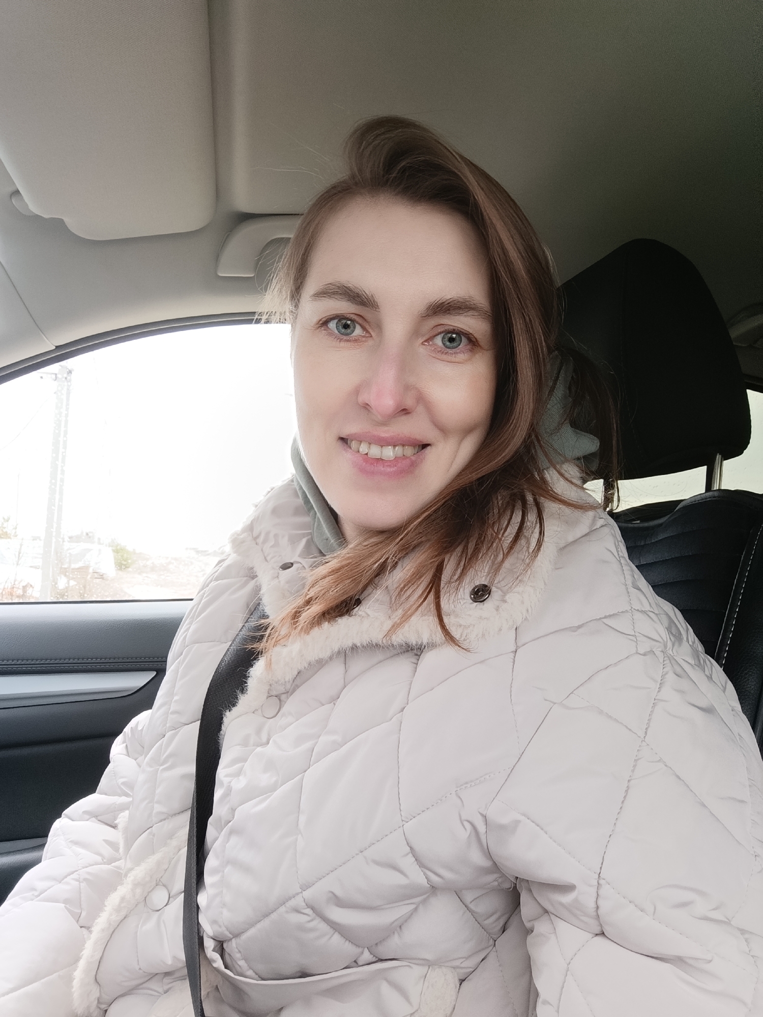 A woman is sitting in the driver's seat of a car. She is wearing a light-colored, quilted jacket with a faux-fur trim. She has brown hair, fair skin, and blue eyes. The woman is wearing a seatbelt and smiling. The car is an SUV with black leather seats and light grey interior trim. The background outside the car window is blurred, but it appears to be a residential area, possibly in Chaikovsky, Russia.