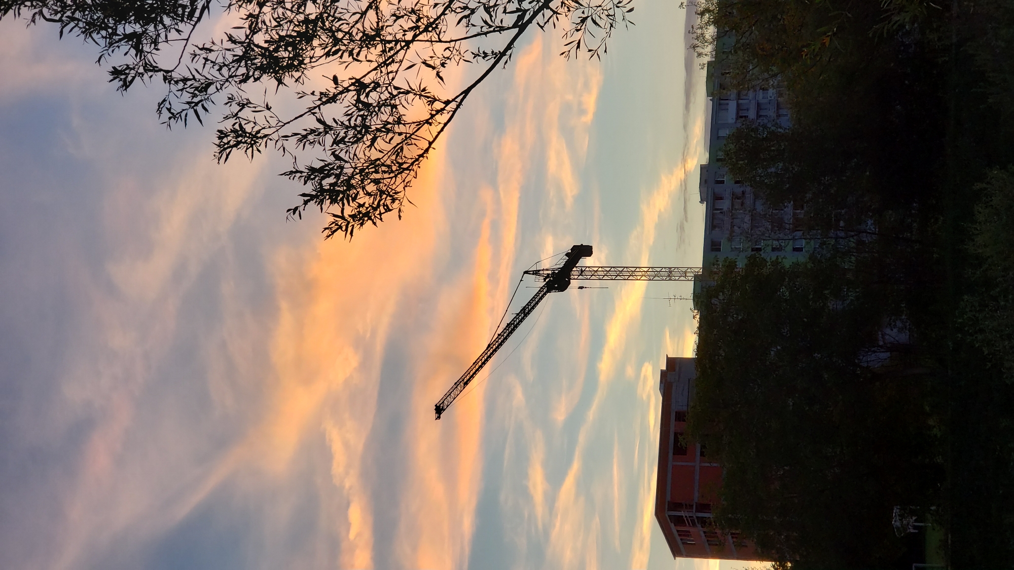 The image depicts a construction site against a backdrop of a sunset sky, with the city of Prešov, Slovakia, serving as the setting. A large construction crane dominates the silhouette against the colorful clouds, which are streaked with hues of orange, pink, and blue. In the foreground, silhouetted trees obscure a portion of the lower part of the frame. Behind the trees, partially visible buildings are under construction or renovation. One building is a reddish-orange structure with visible concrete framework, suggesting ongoing work. Adjacent to it, a taller, multi-story building with a light green facade stands, appearing to be an apartment complex. The overall scene suggests a typical urban evening at a location where development is taking place. There are no people or discernible actions captured in this still image.