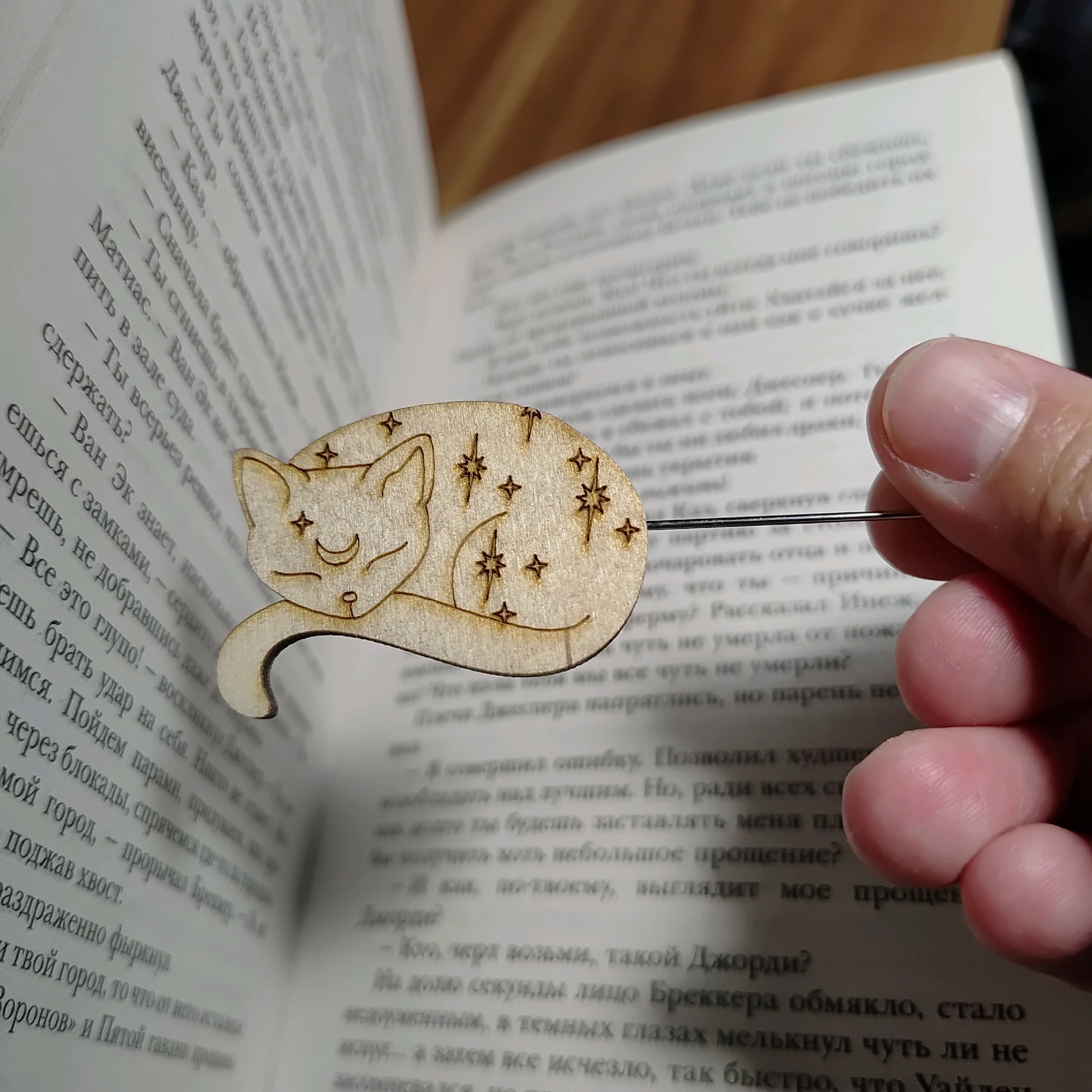 The image shows a wooden bookmark shaped like a sleeping cat with stars and a crescent moon on it, inserted into an open book. The bookmark is held by a person's fingers, which are visible on the right side of the frame. The book's pages are filled with text in Russian. The background is blurred, but appears to be a wooden surface, possibly a table or desk. There is no discernible information in the image that would allow for the determination of the location within the city of Марковский, Russia.