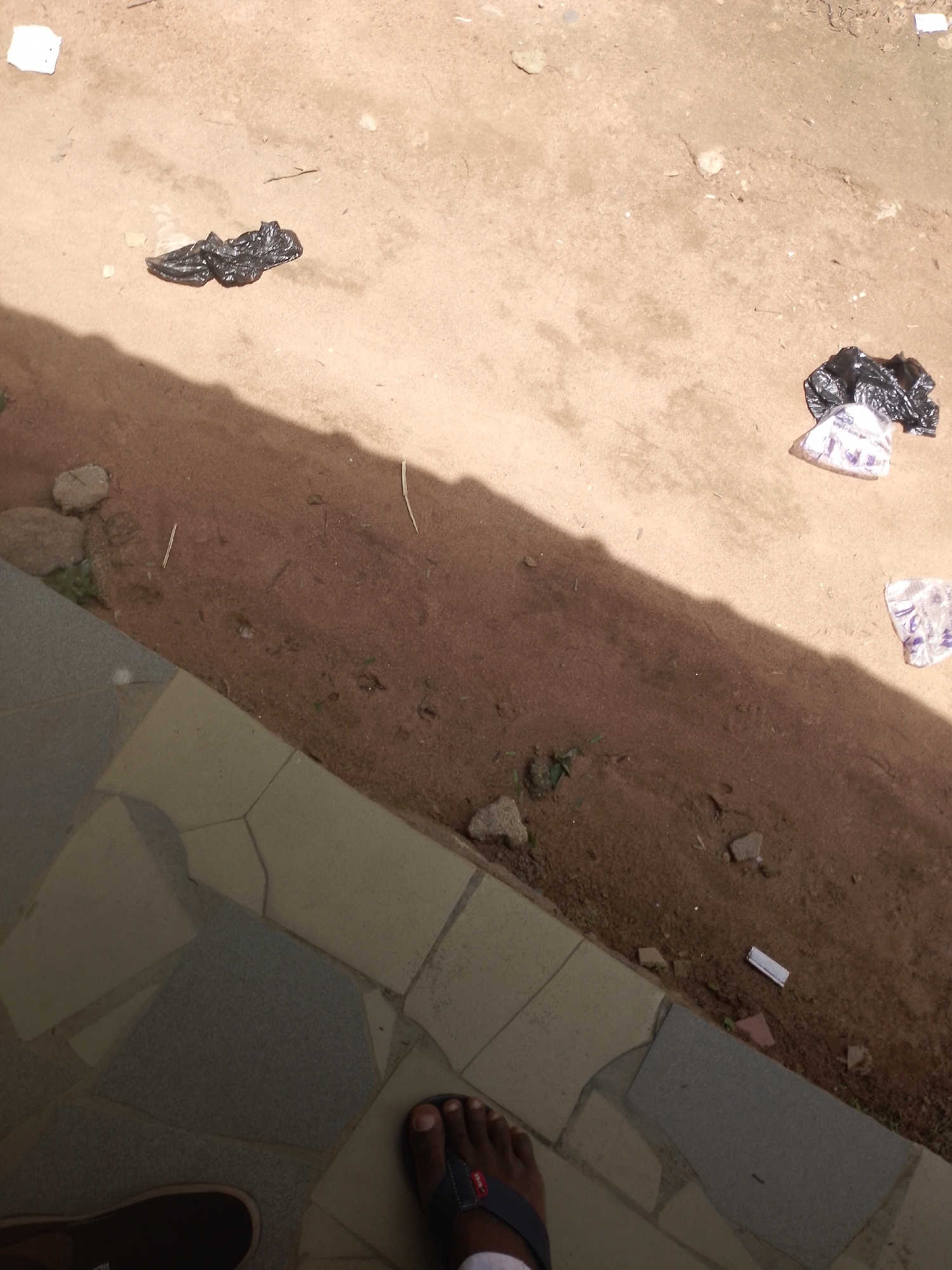 A high-angle outdoor shot shows a person's feet wearing dark sandals and brown shoes on a tiled surface. The ground beyond the tiles is sandy dirt with some small rocks and scattered debris, including black plastic bags and a white plastic bag. A shadow falls across the sandy area from the left side of the frame. The scene is devoid of any identifiable landmarks or specific contextual clues that would indicate a precise location within a city.