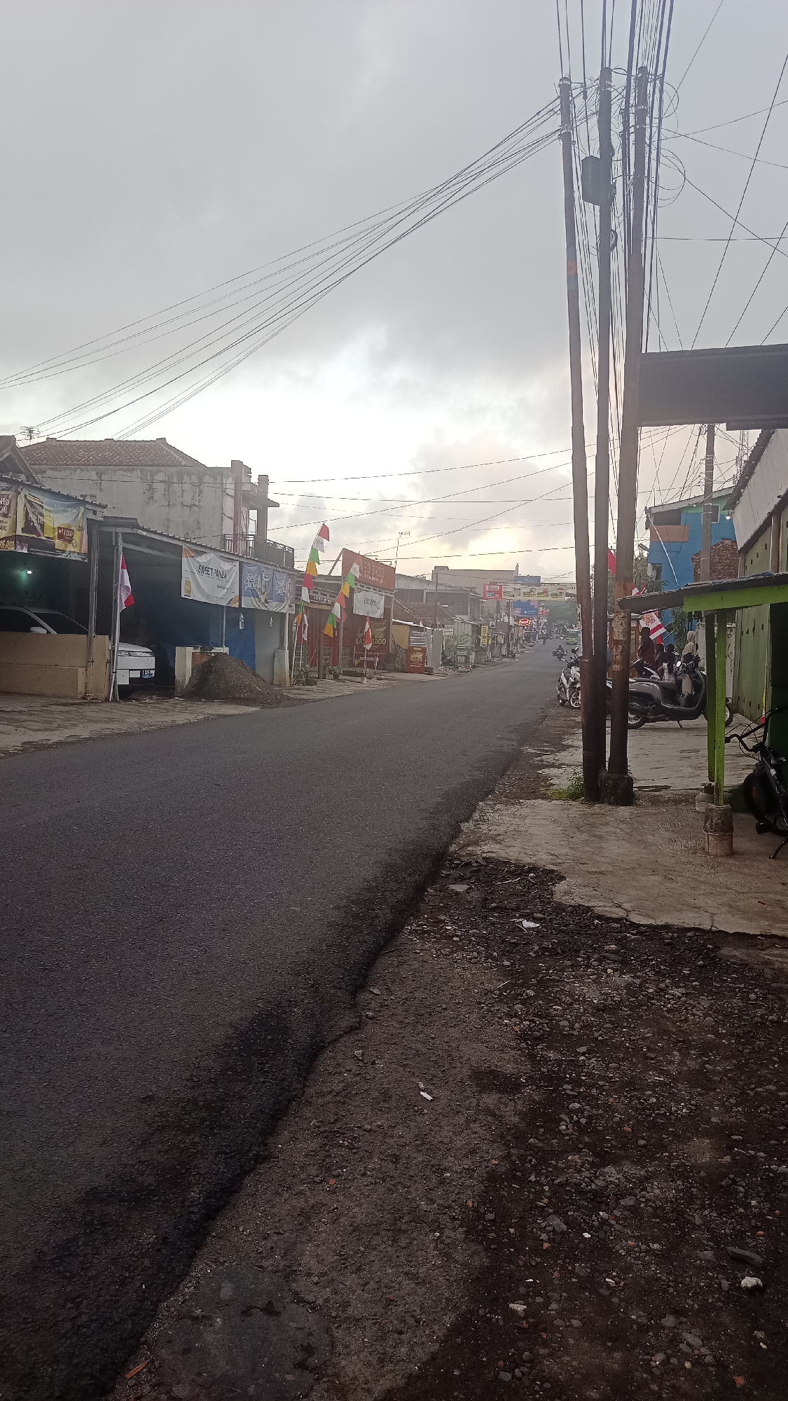 This is a street-level photograph taken in Pakemitan, Indonesia, depicting a rural or suburban street scene. The time of day appears to be early morning or late afternoon due to the overcast sky and diffused lighting. The road is paved with asphalt and shows some signs of wear and tear. On the left side of the road, there are several buildings that appear to be shops or small businesses, some with awnings and Indonesian flags displayed. A white car is parked in front of one of these establishments. On the right side of the road, there are more buildings, including one with a green awning. Several motorcycles are parked on the sidewalk in front of these buildings. Overhead, a complex network of power lines crisscrosses the sky, supported by wooden utility poles. The overall impression is of a quiet, functional street in a developing area.