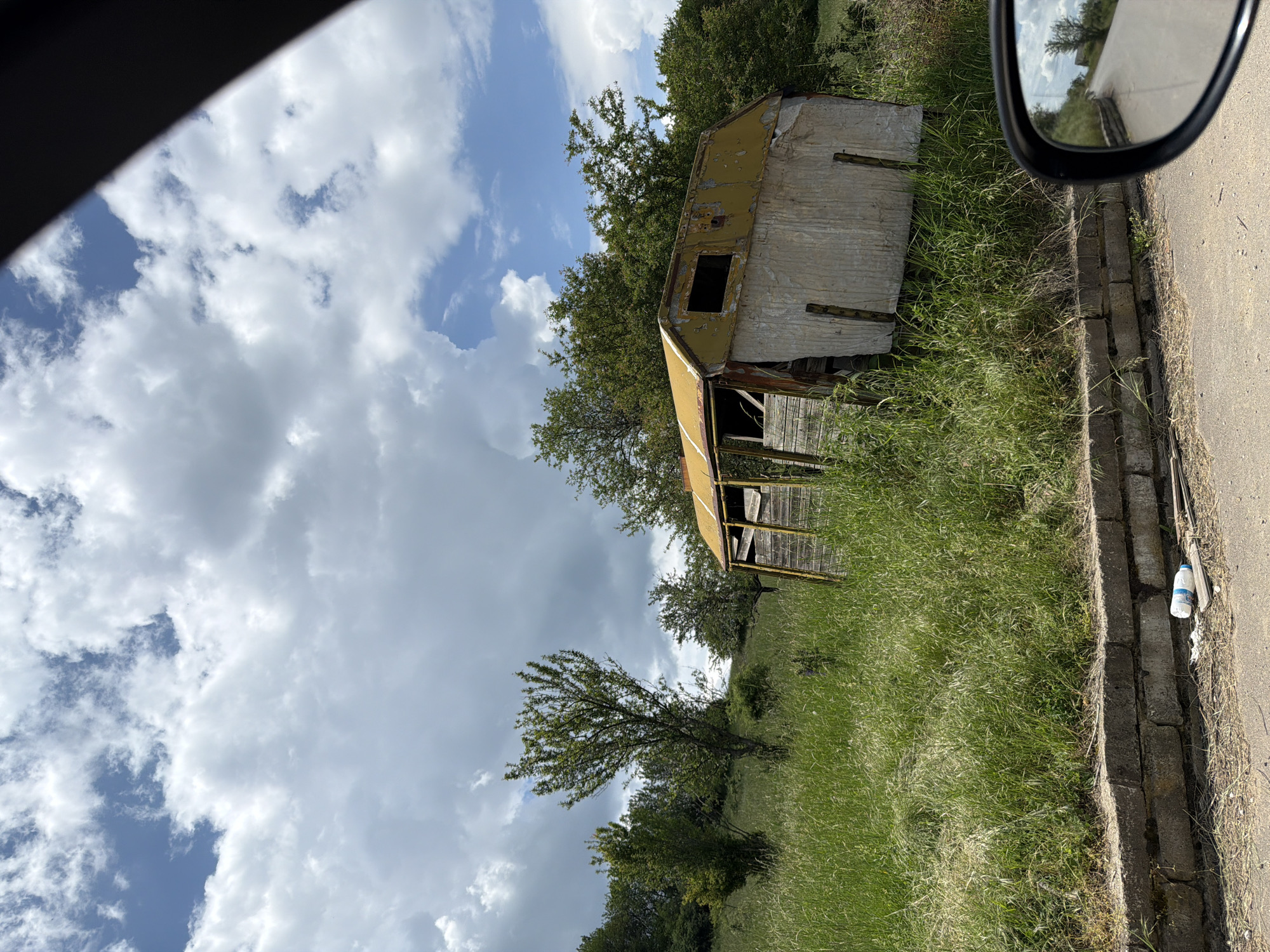 An old, derelict structure sits in a grassy field. The structure is primarily constructed of metal sheets, some painted yellow, with a simple frame visible through openings. A tree grows near the structure, partially obscuring it. The sky is filled with clouds. A road borders the field, and a side-view mirror is visible in the upper right corner. A white plastic bottle rests on the road's edge. It is impossible to confirm the location in Gurgulyat, Bulgaria without additional information.